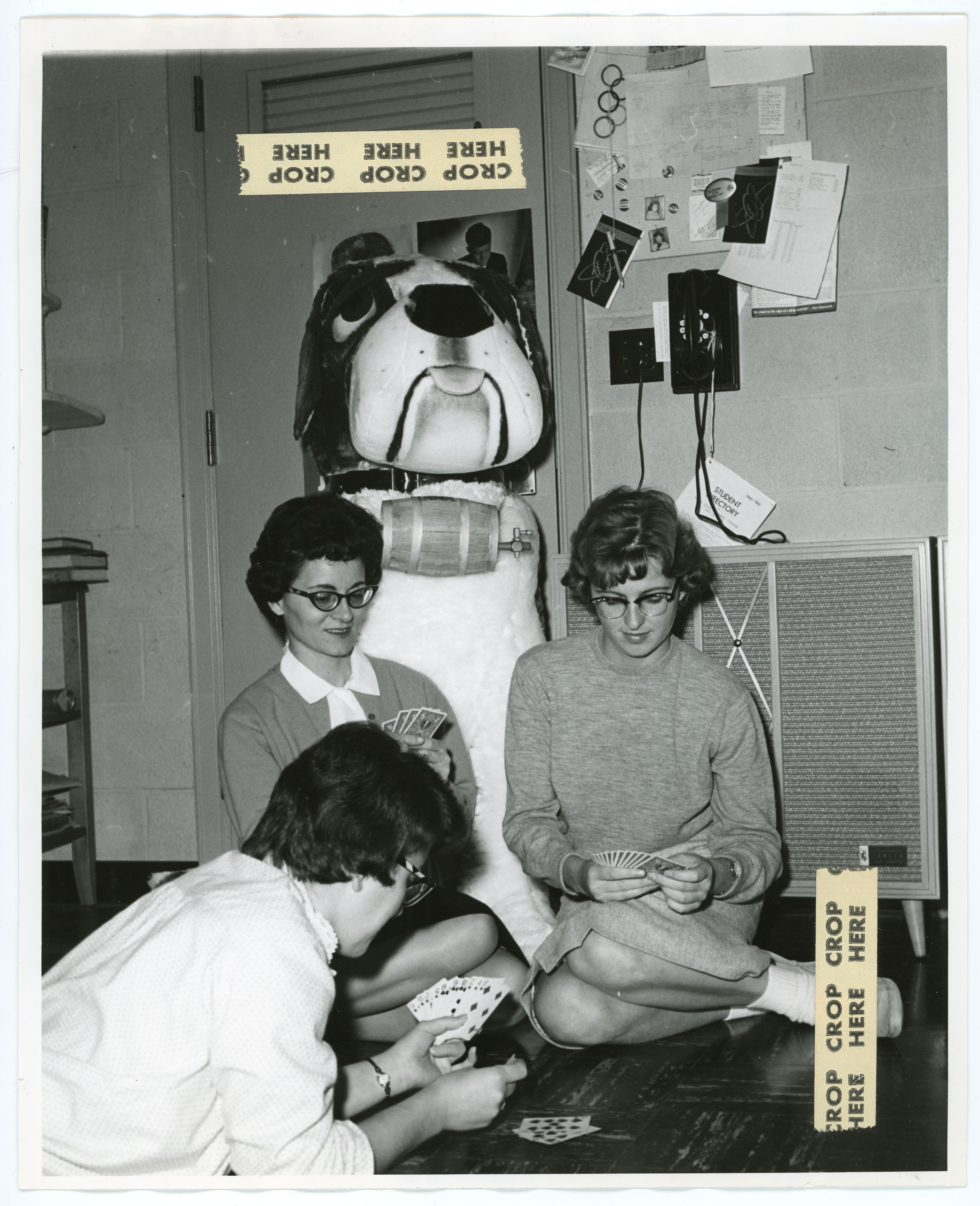 Three women playing cards in dorm room with large statue of dog behind them