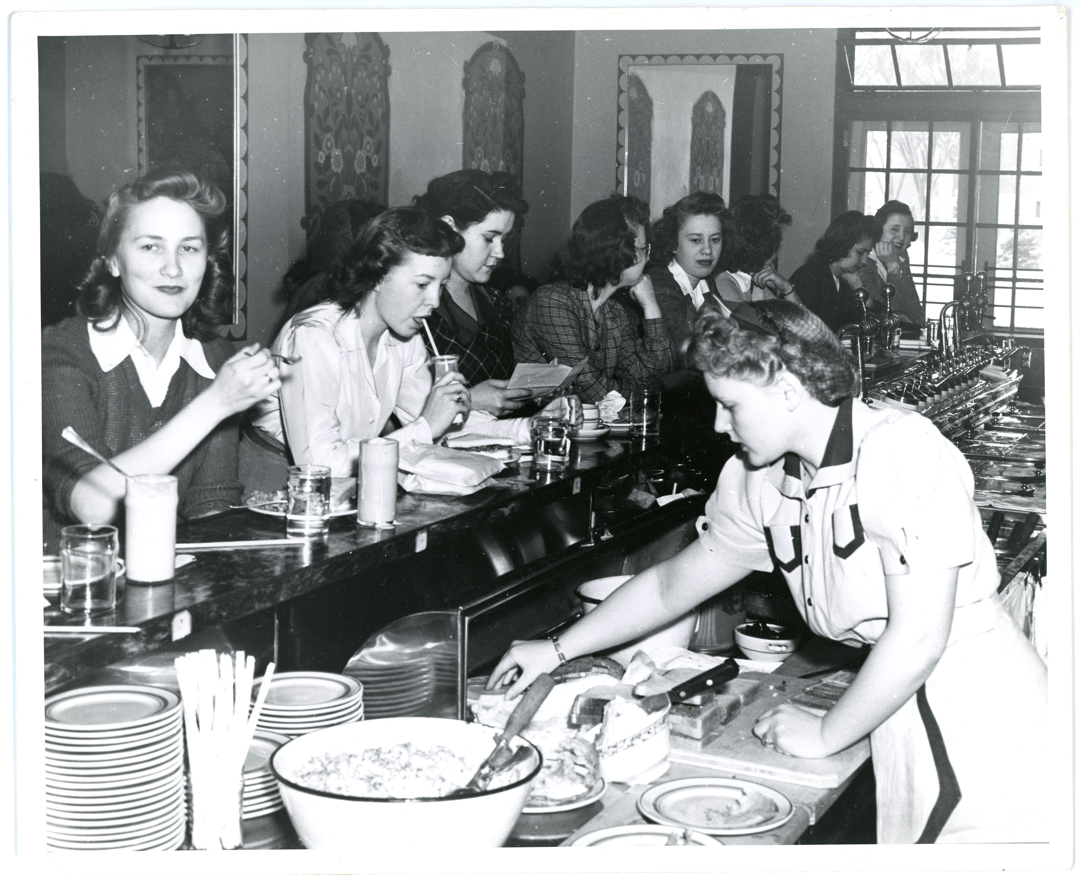 One woman serving eight women seated at a soda fountain counter with food and drinks