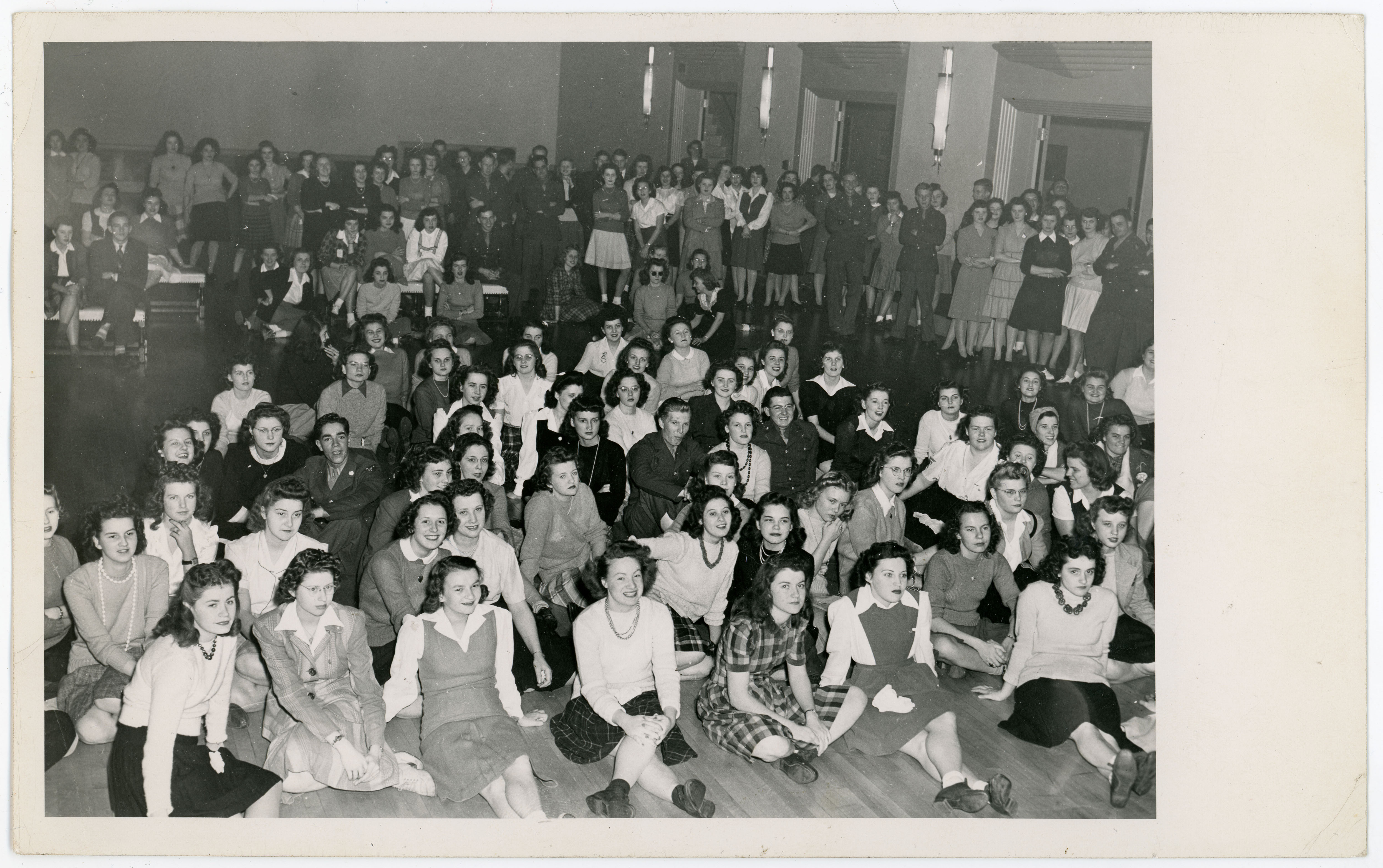 Crowd of people gathered in a ballroom, seated on the floor and bleachers as well as standing, to watch an off-camera program