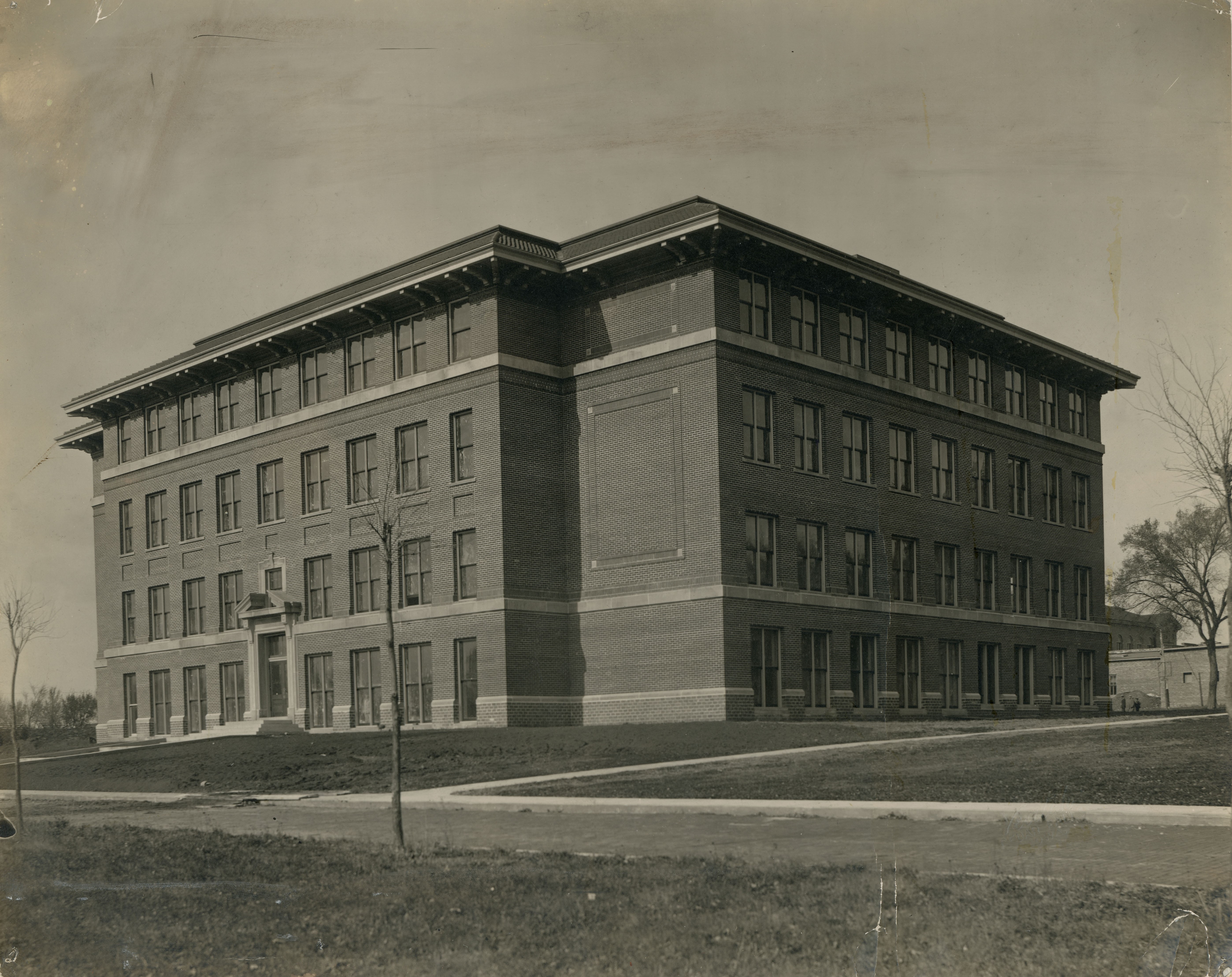 Four-story brick building with windows on each level, two sides visible, bare dirt and short trees surrounding