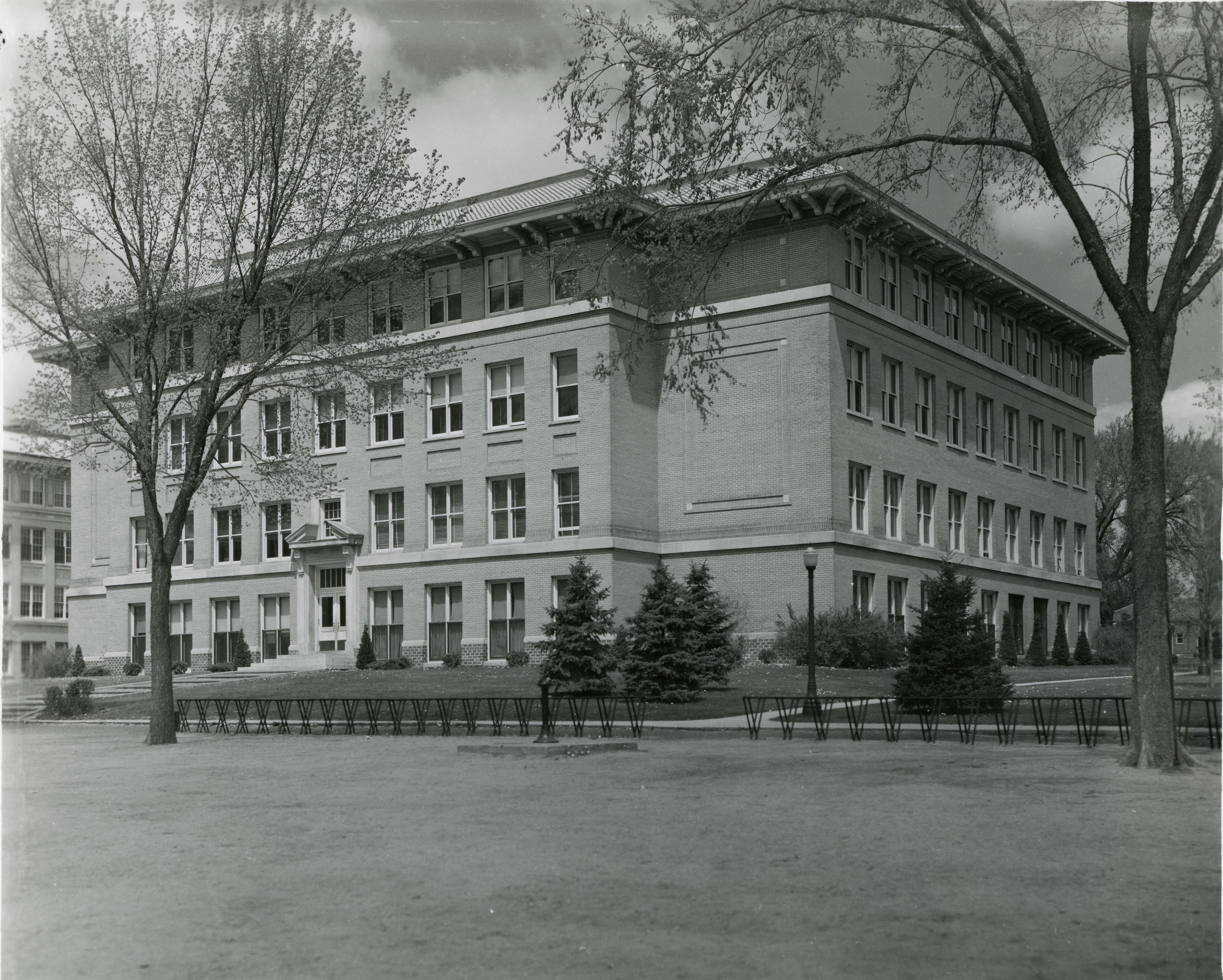 Four-story brick building with windows on each level, two sides visible, bike racks and trees in front of it