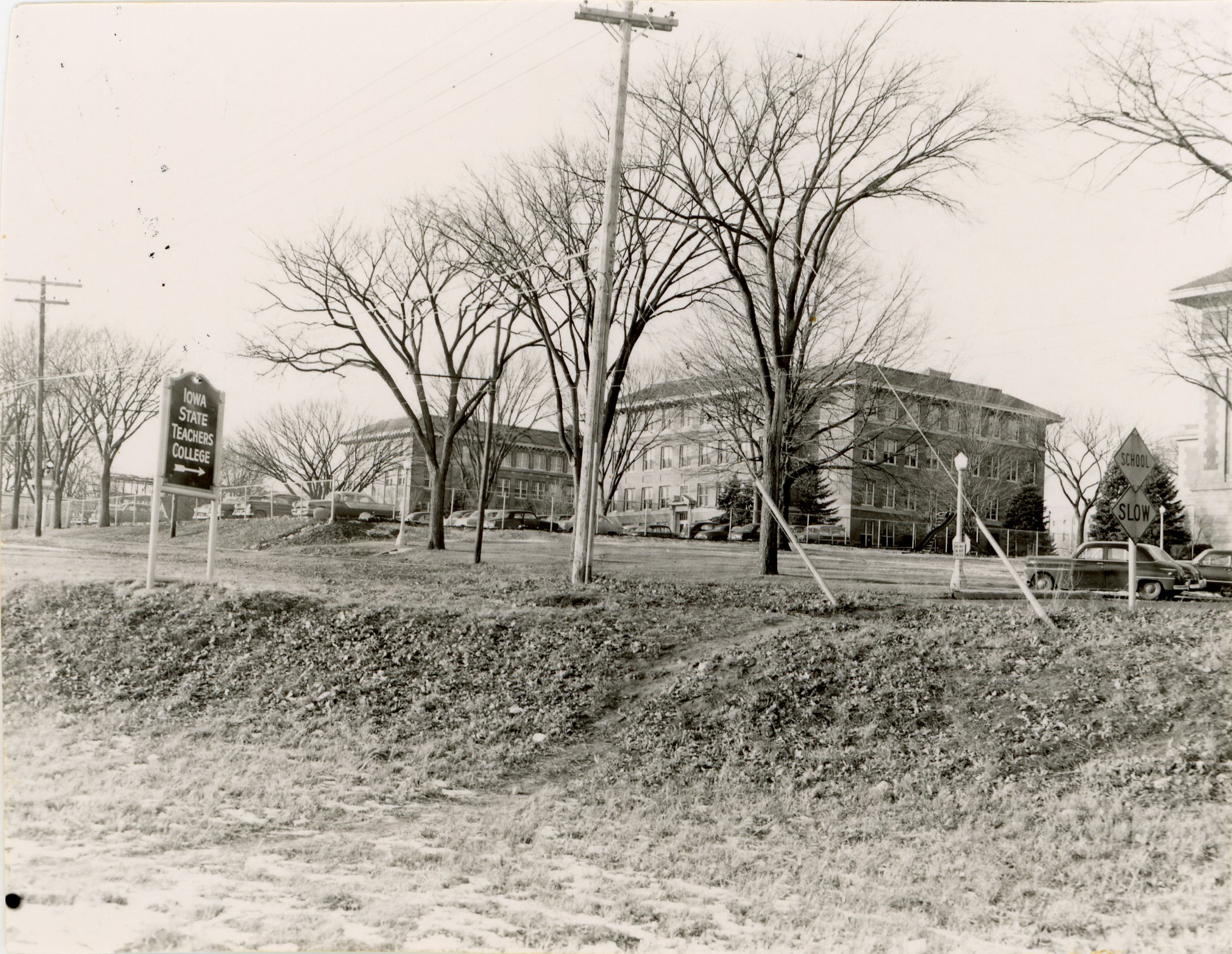 A sign reading Iowa State Teachers College with arrow pointing toward three visible buildings with cars parked in front of them