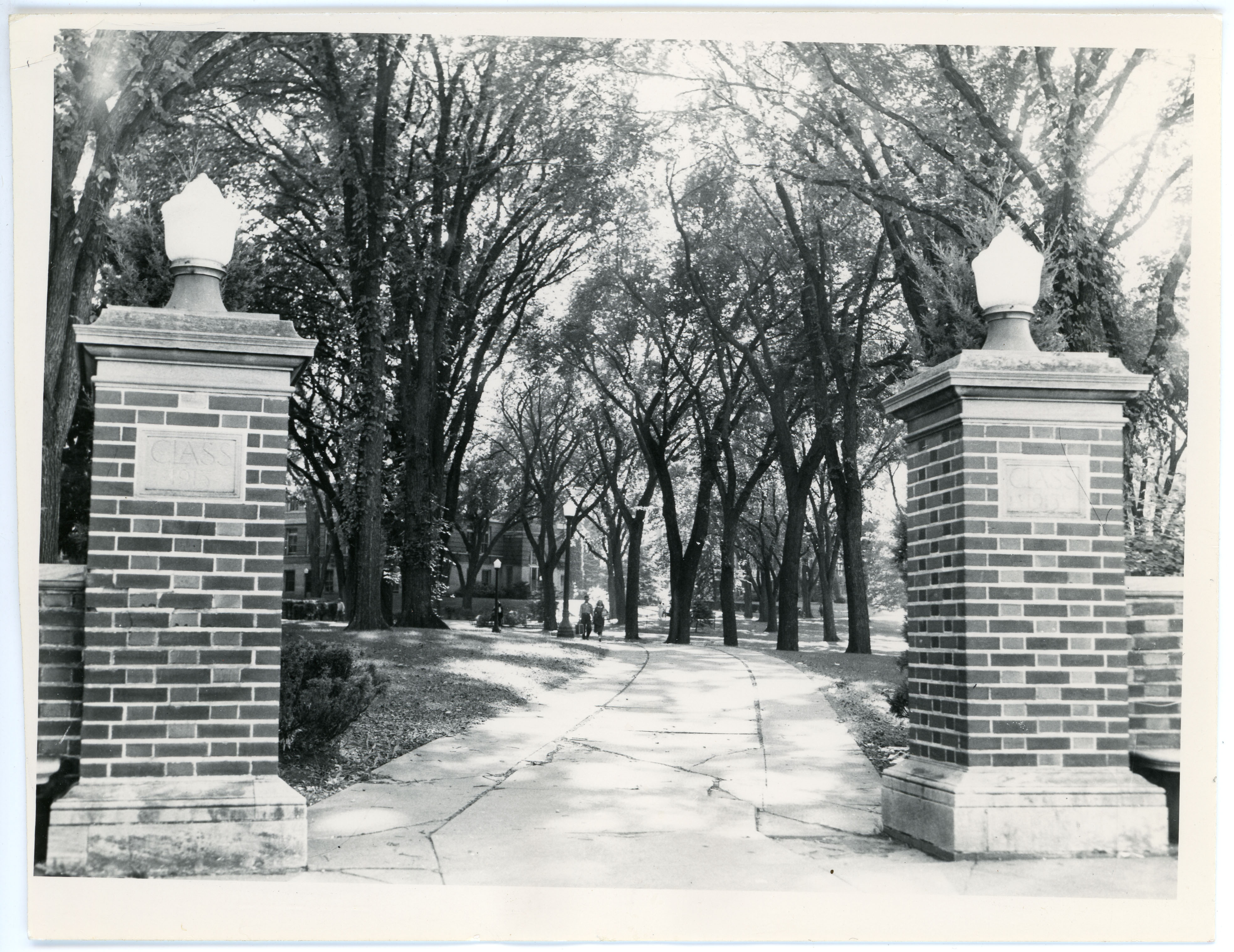 Two brick pillars on either side of a concrete path with trees in the background