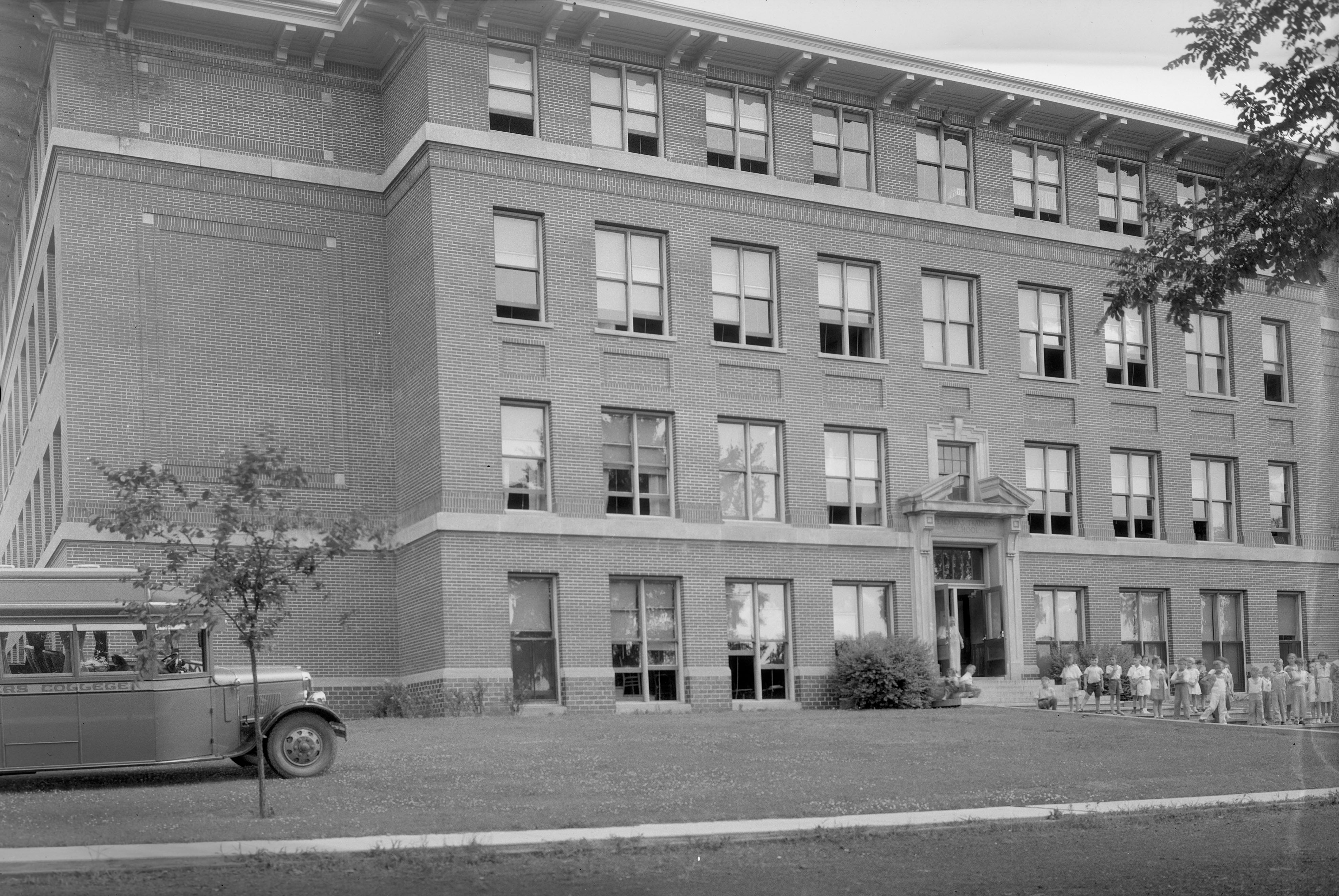 A bus parked on the left side of a four-story brick building with a line of children on the right standing before the building entrance