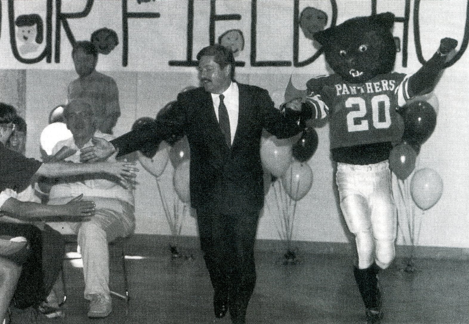 Man in suit holding hands with panther mascot to right and giving high-fives to people on left, balloons in background