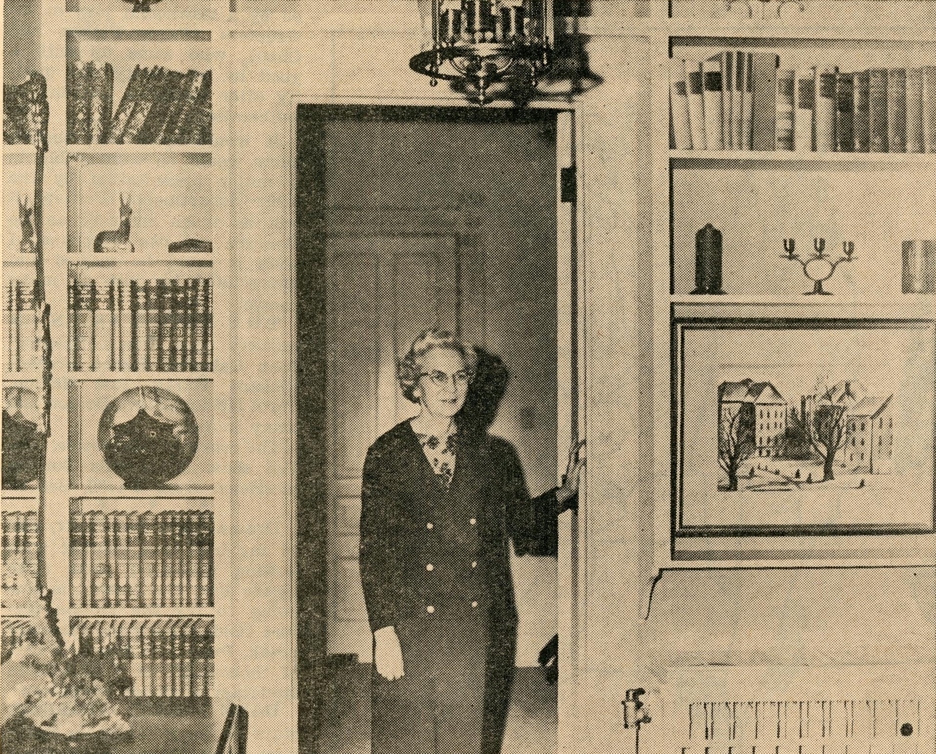 Printed in newspaper, woman standing in an open interior of a house doorway which is framed by built-in shelves