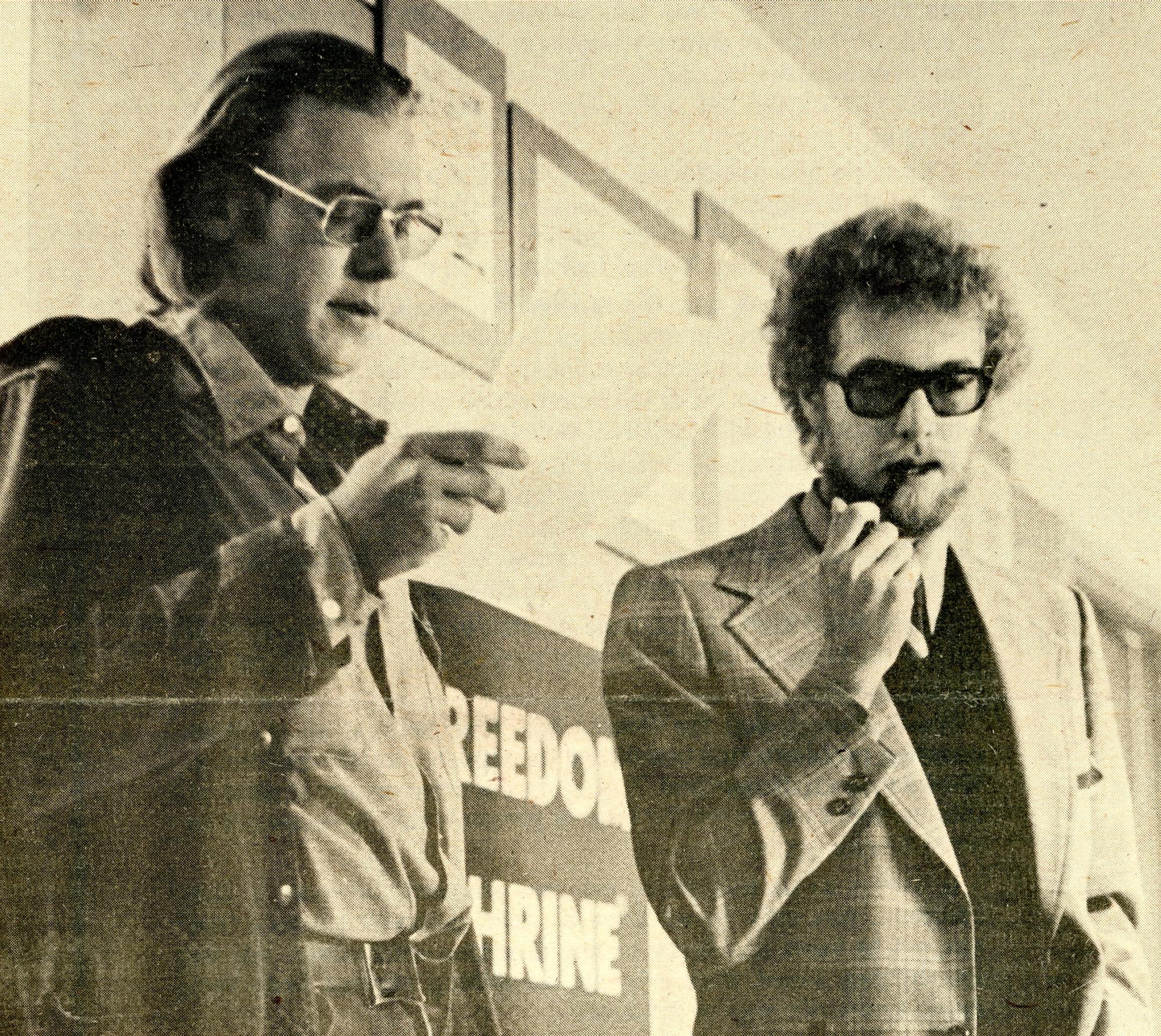 Two men in a hallway with signs mounted on the wall; man on left is reading a paper and man on right is smoking a pipe