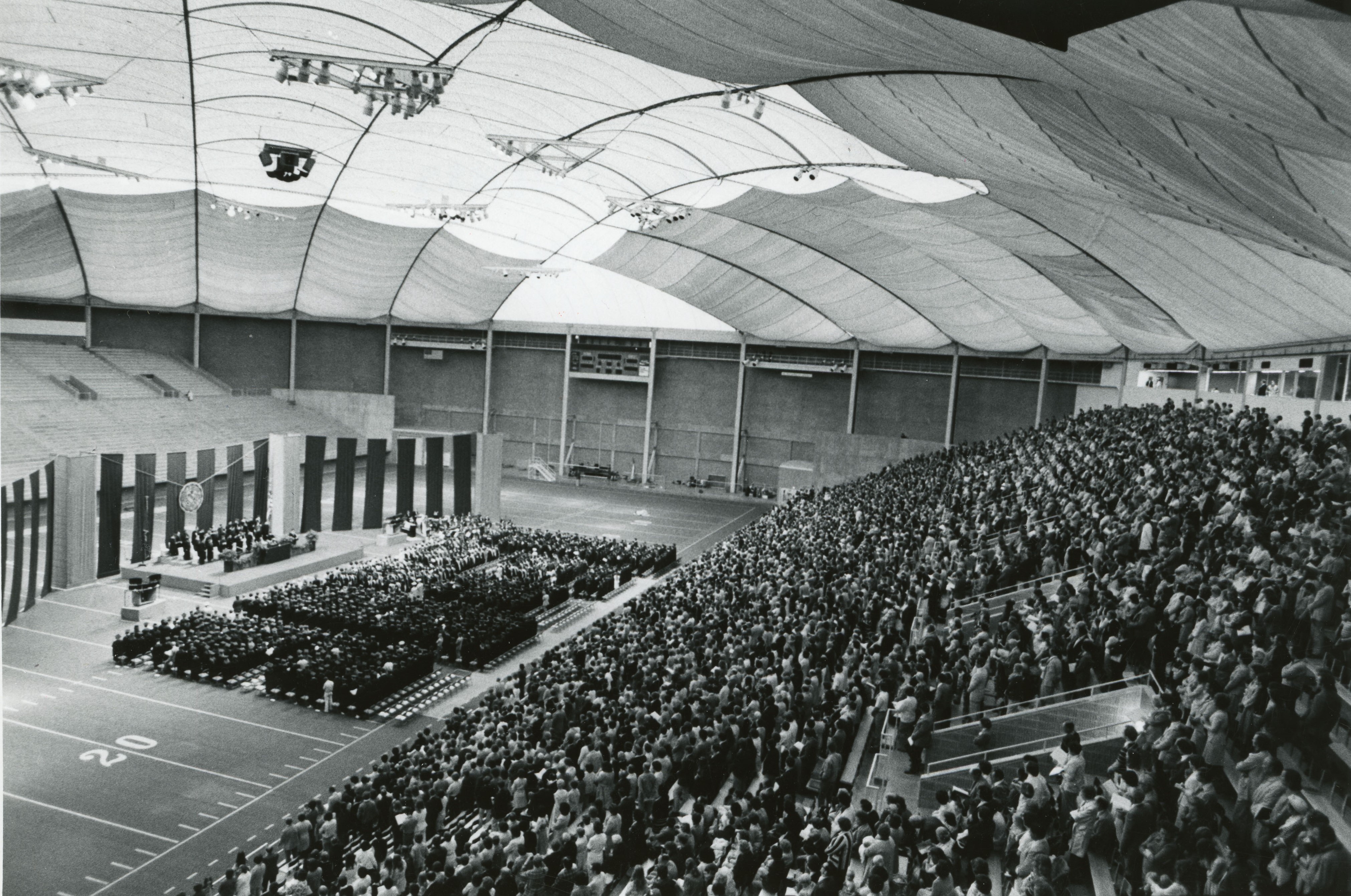 Stage set up for commencement and graduates on field to left side, crowd filling seats along right