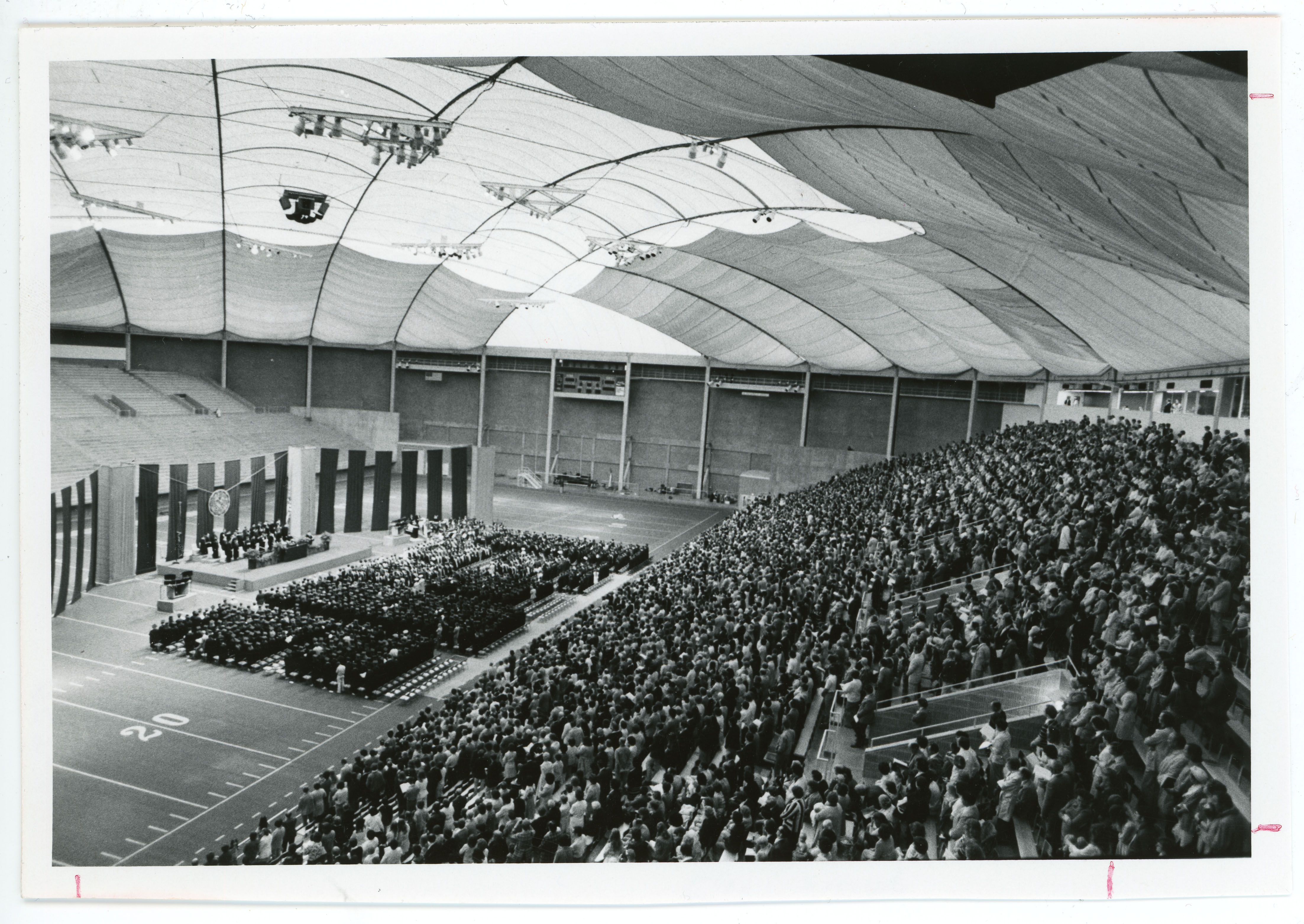 Stage set up for commencement and graduates on field to left side, crowd filling seats along right