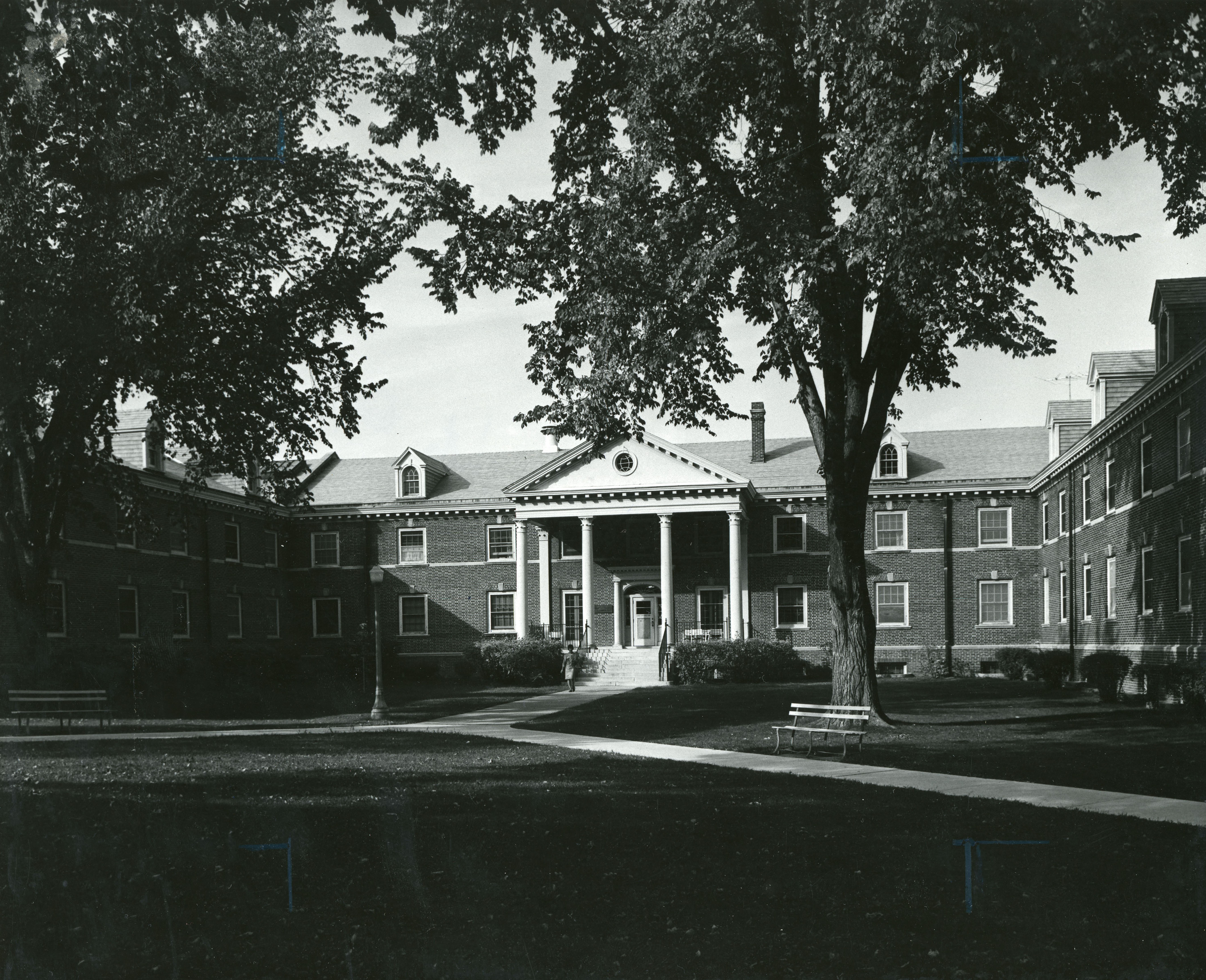 Two-story C-shaped brick building with pillared covered entrance, framed by trees and lawn