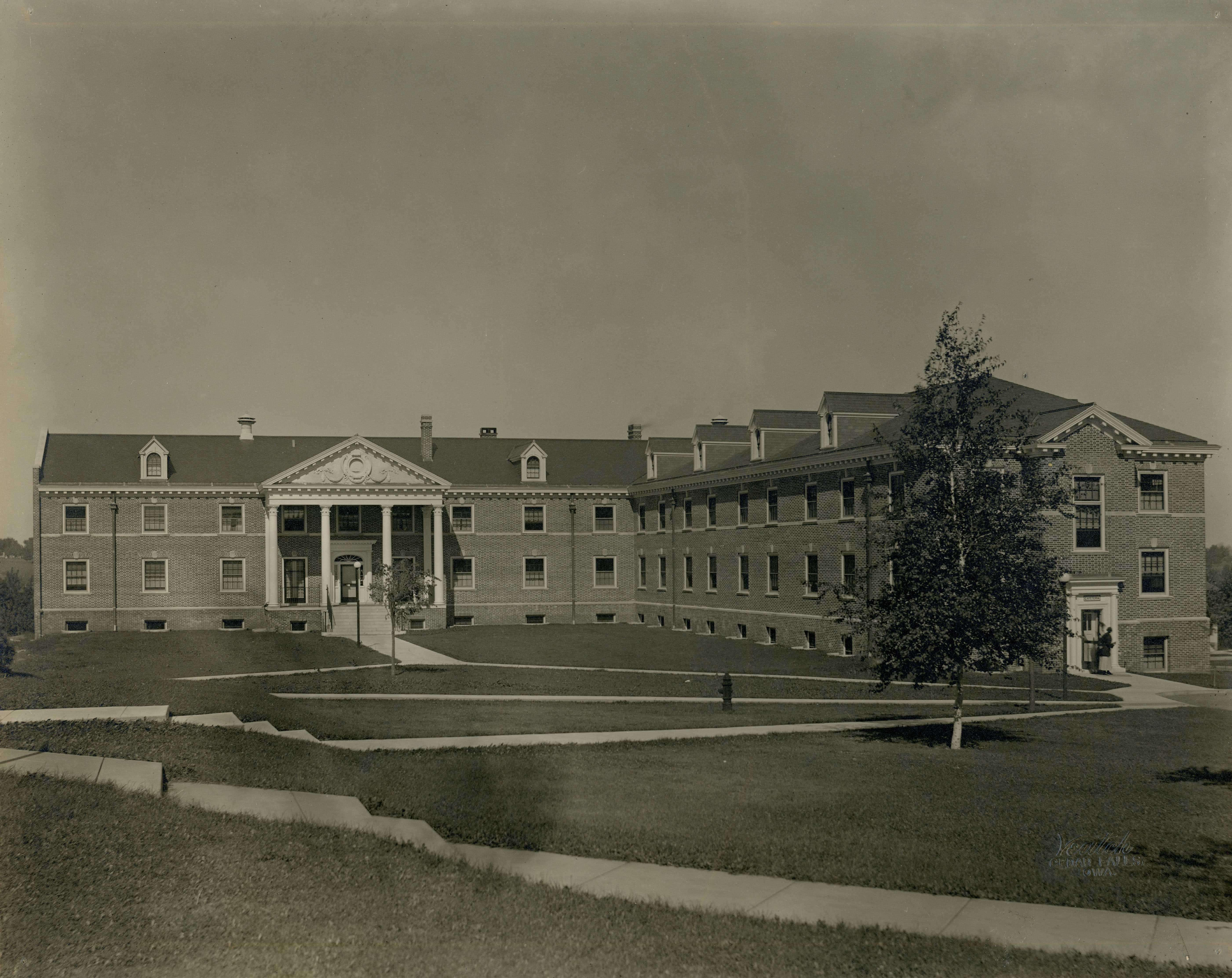 L-shaped two-story brick building with pillared covered entrance shot across a lawn