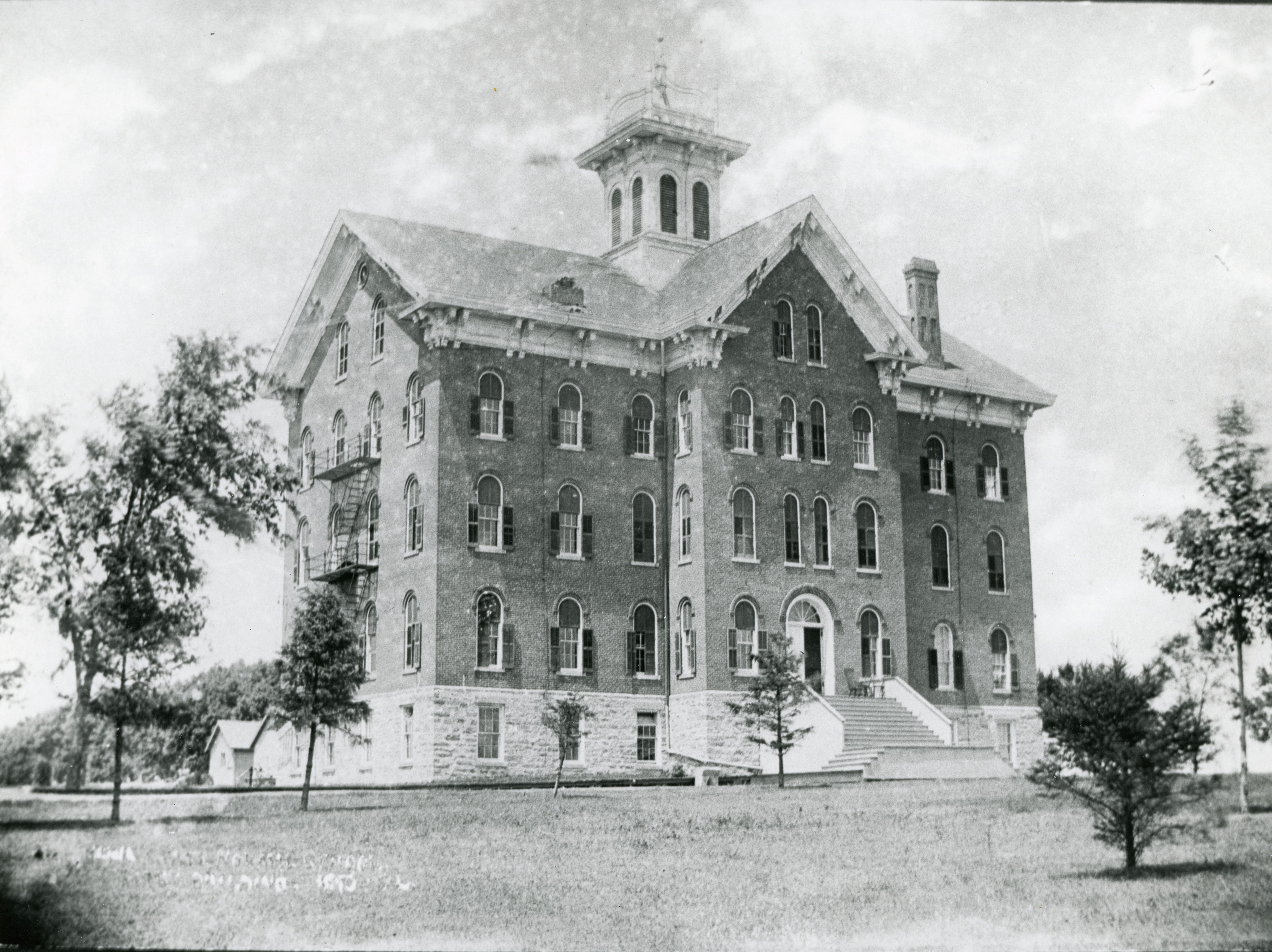 Three and a half story brick and stone building with a cupola; caption in lower left is illegible