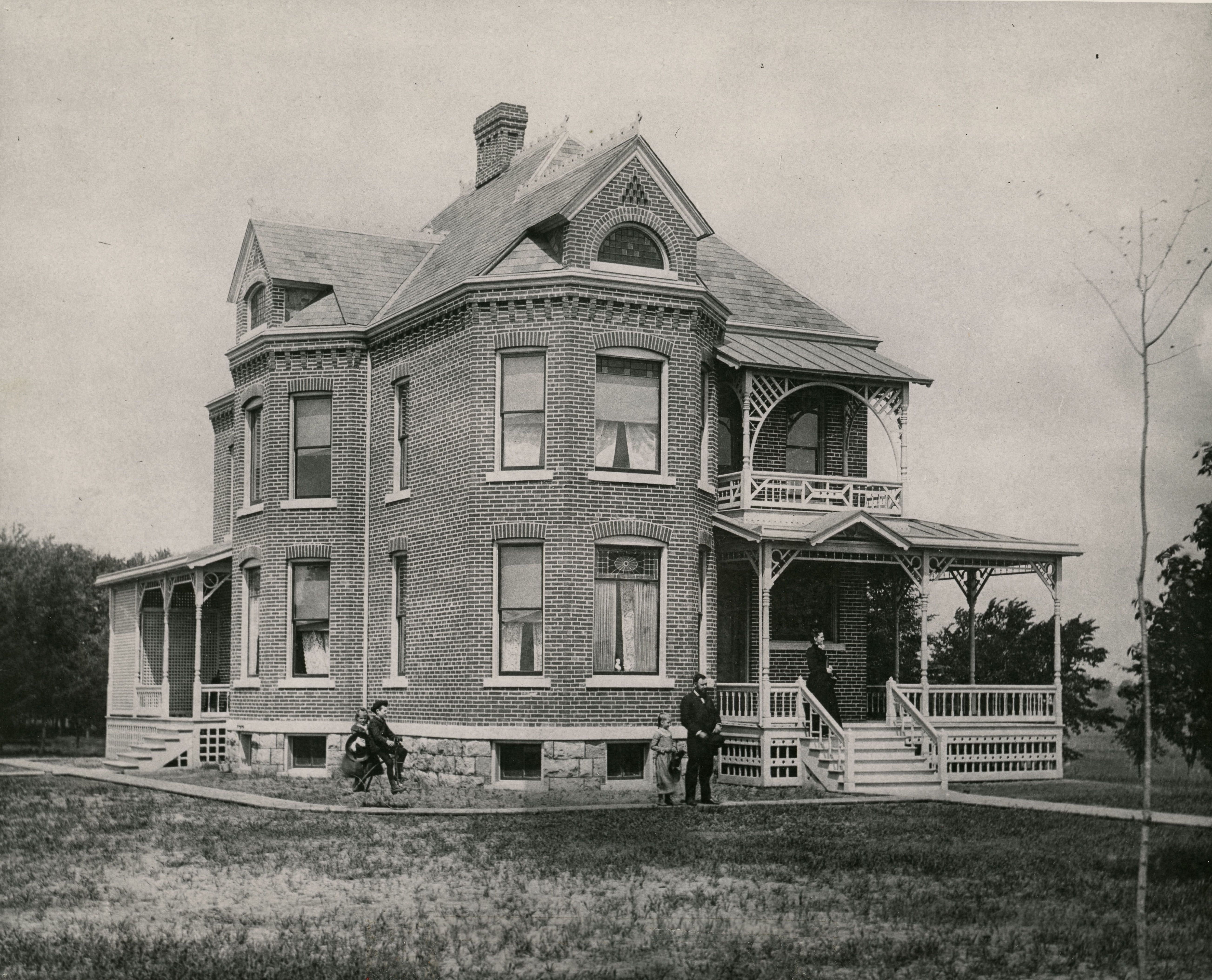 Two and a half story brick house with two porches on visible sides and one balcony, a woman standing on the front porch and a man and three children standing alongside the house