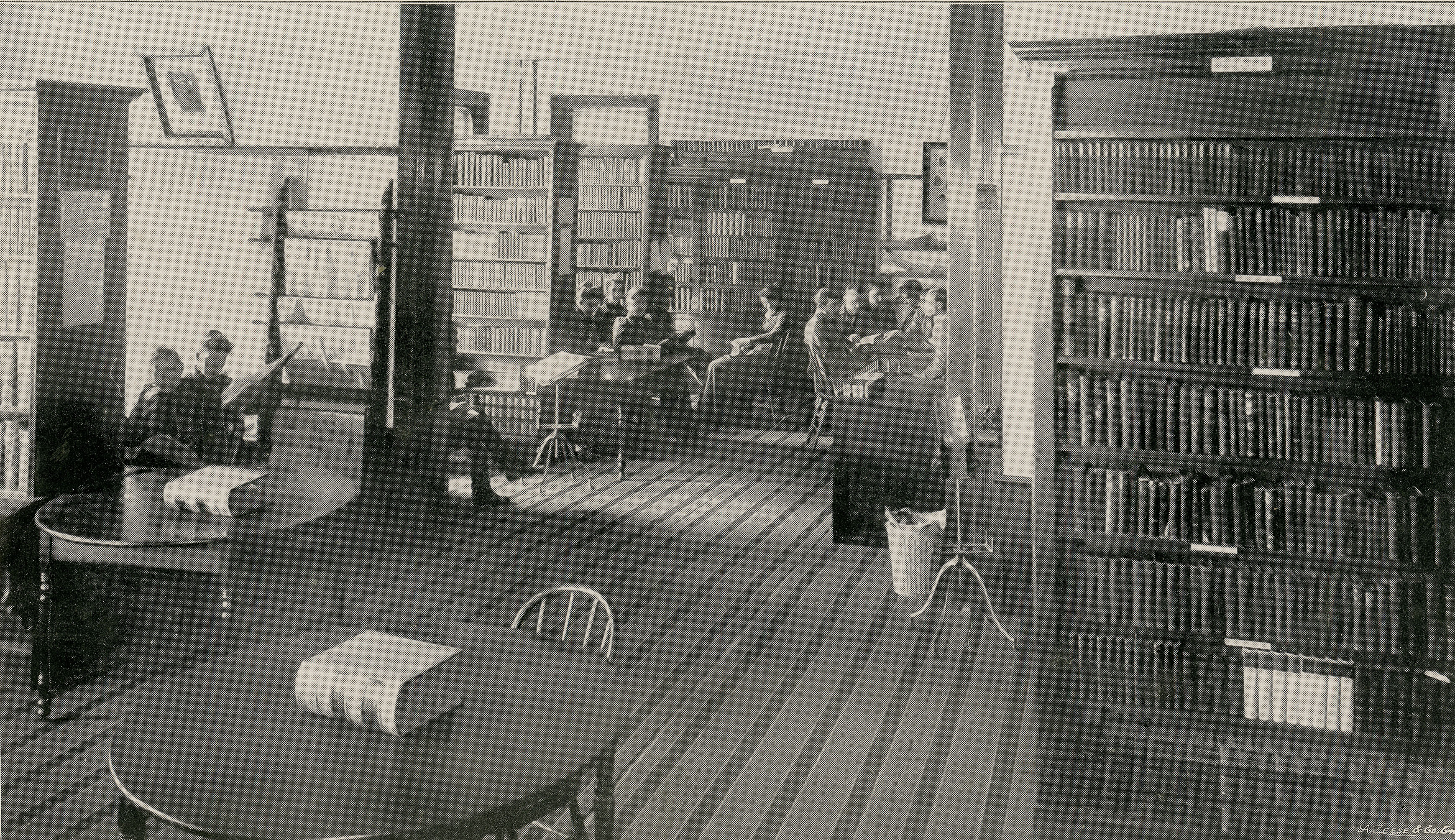 Students seated around tables among library stacks in a room with striped carpet