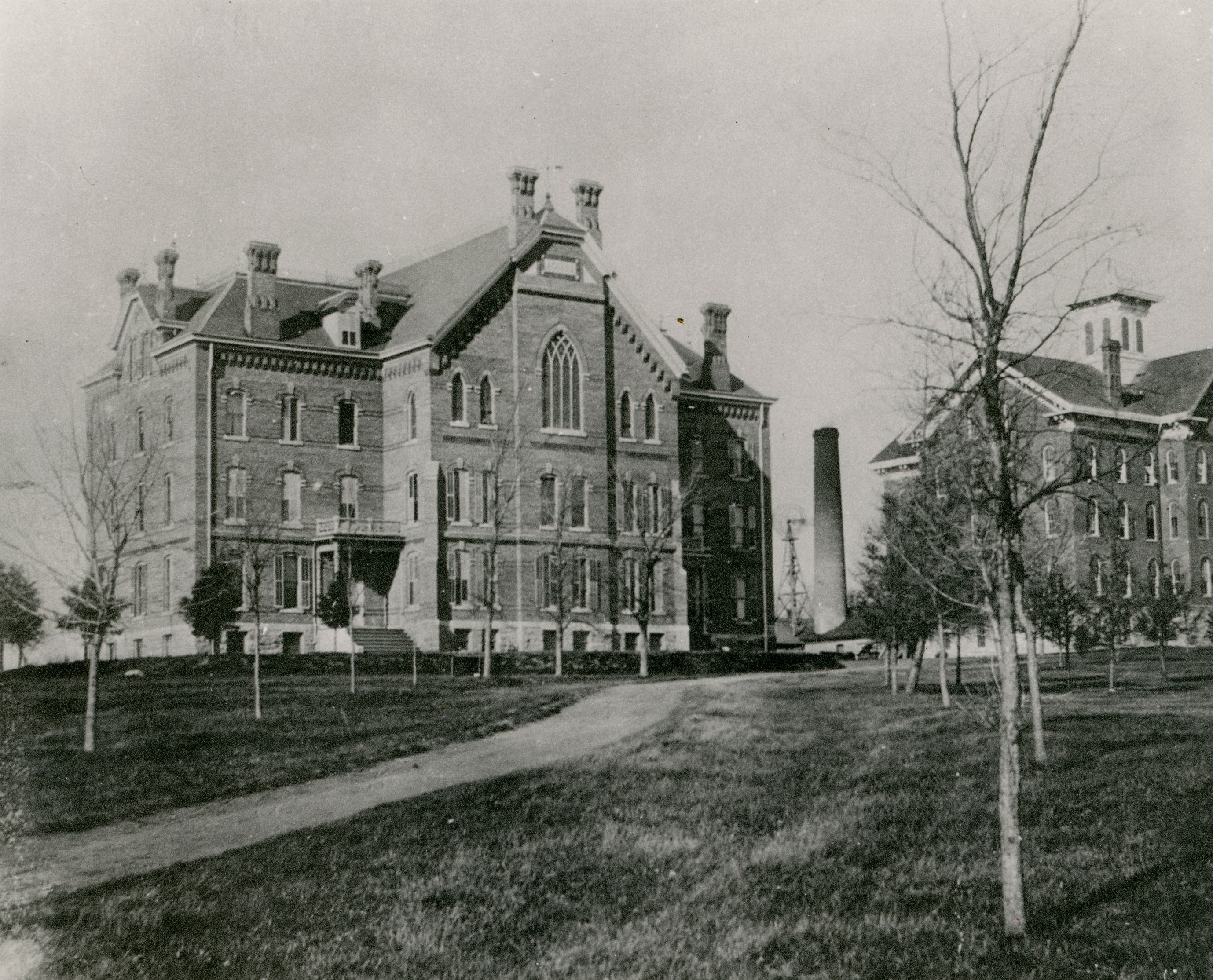 Three and a half story brick building with pointed arched window on upper floor to left and a three and a half story brick building with cupola to the right; visible between are a windmill and smokestack