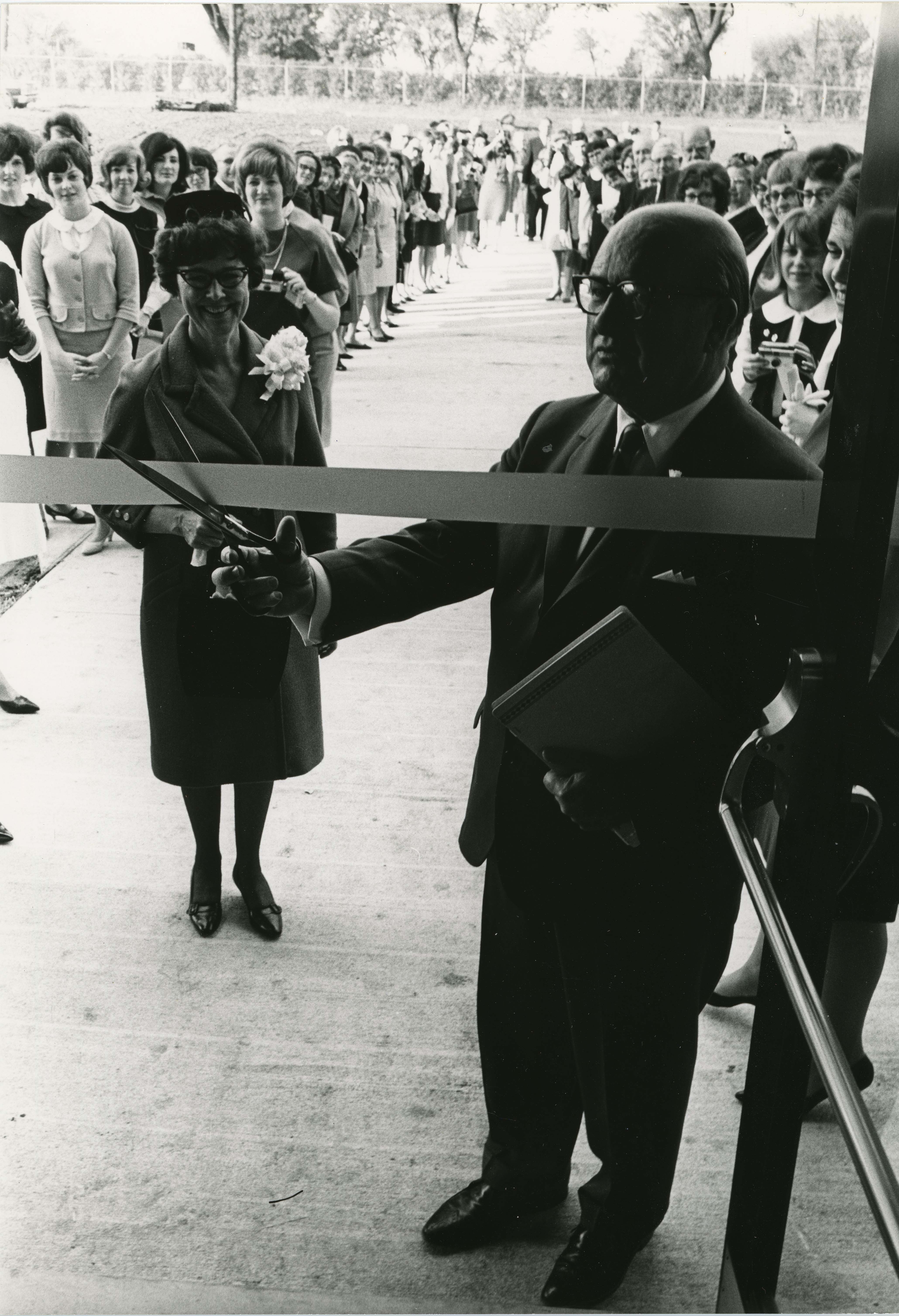 A man cutting a ribbon across a doorway, crowd in background
