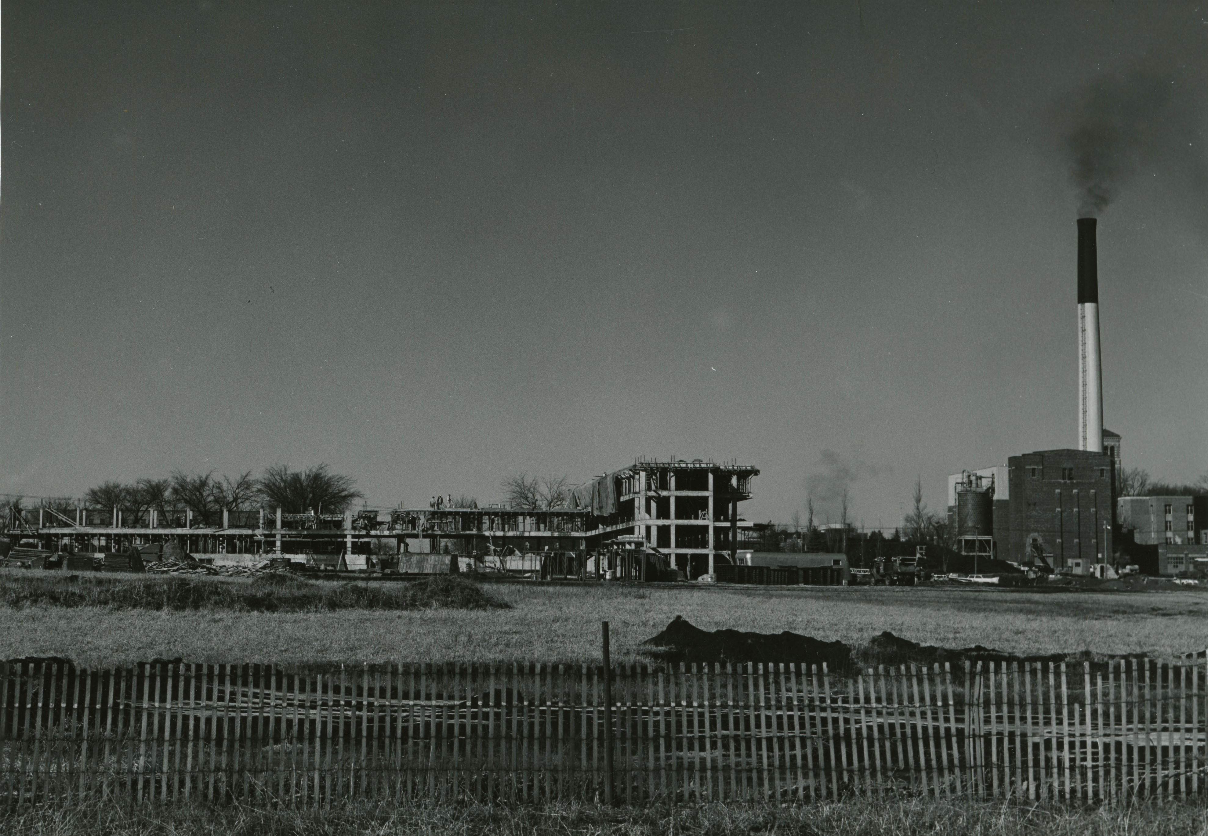Building under construction with fencing in front of the work area field and a smokestack and powerpoint to the right