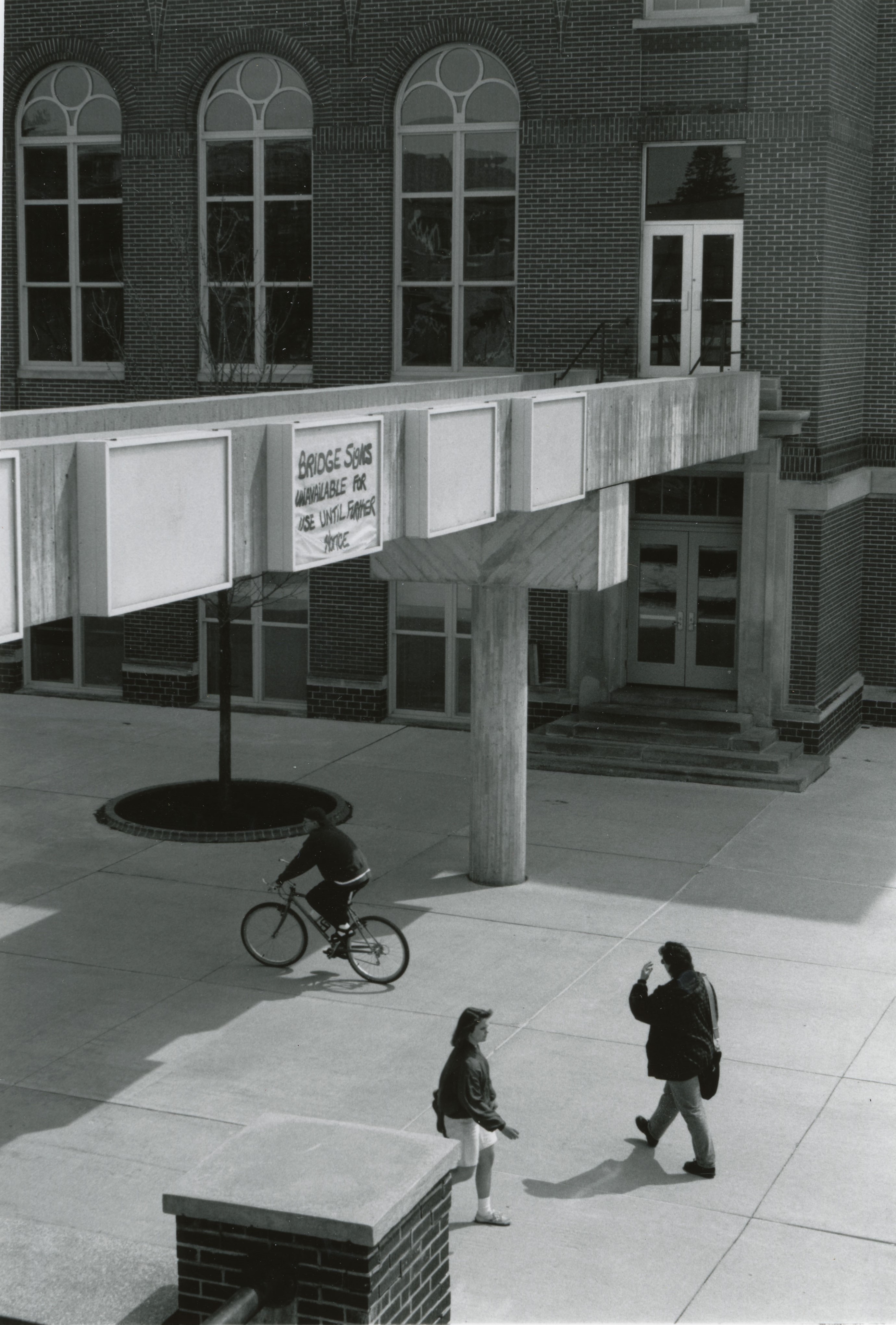 One person biking and two walking under concrete bridge between buildings with sign on it reading: Bridge signs unavailable for use until further notice