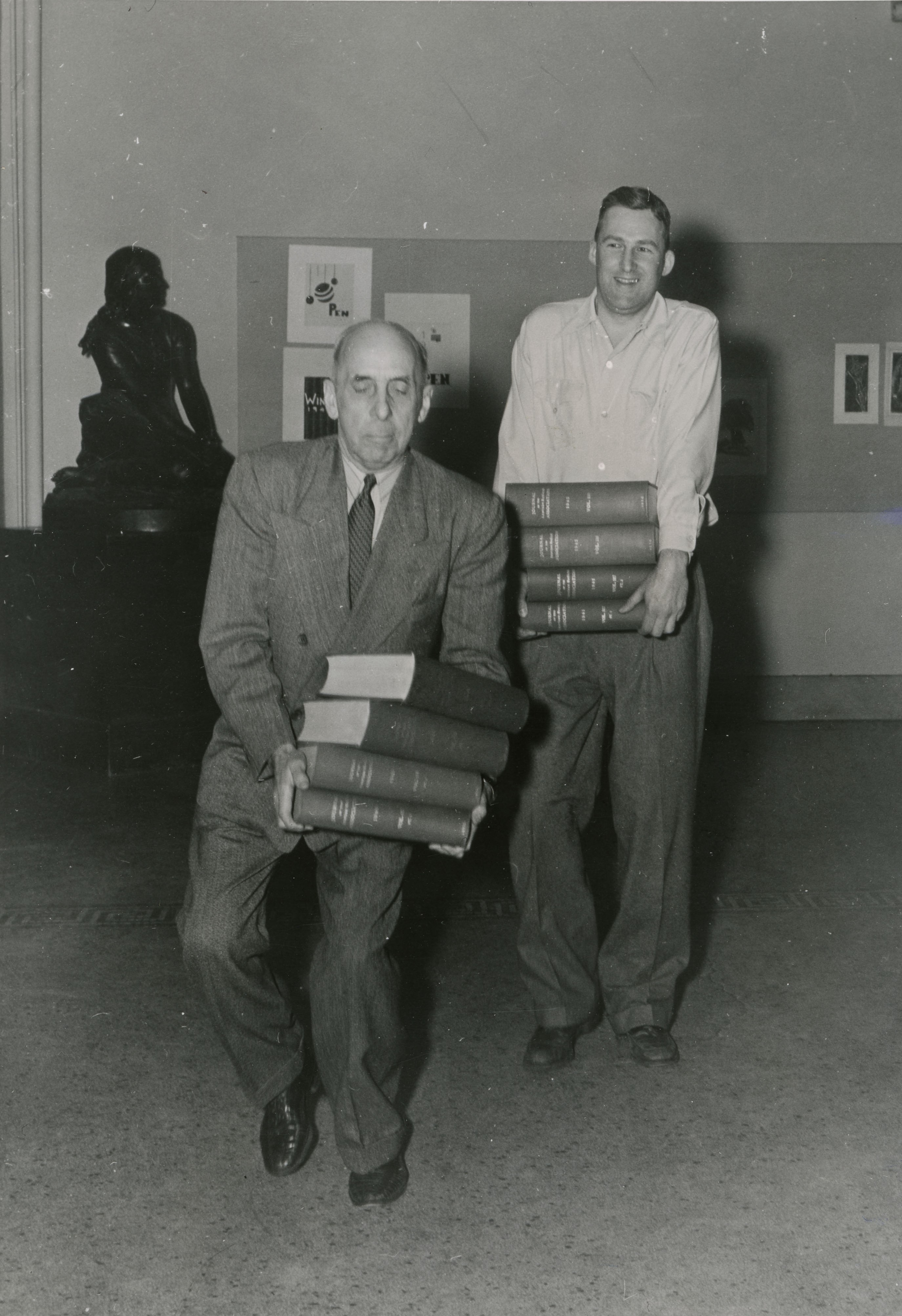 Two men precariously carrying stacks of books