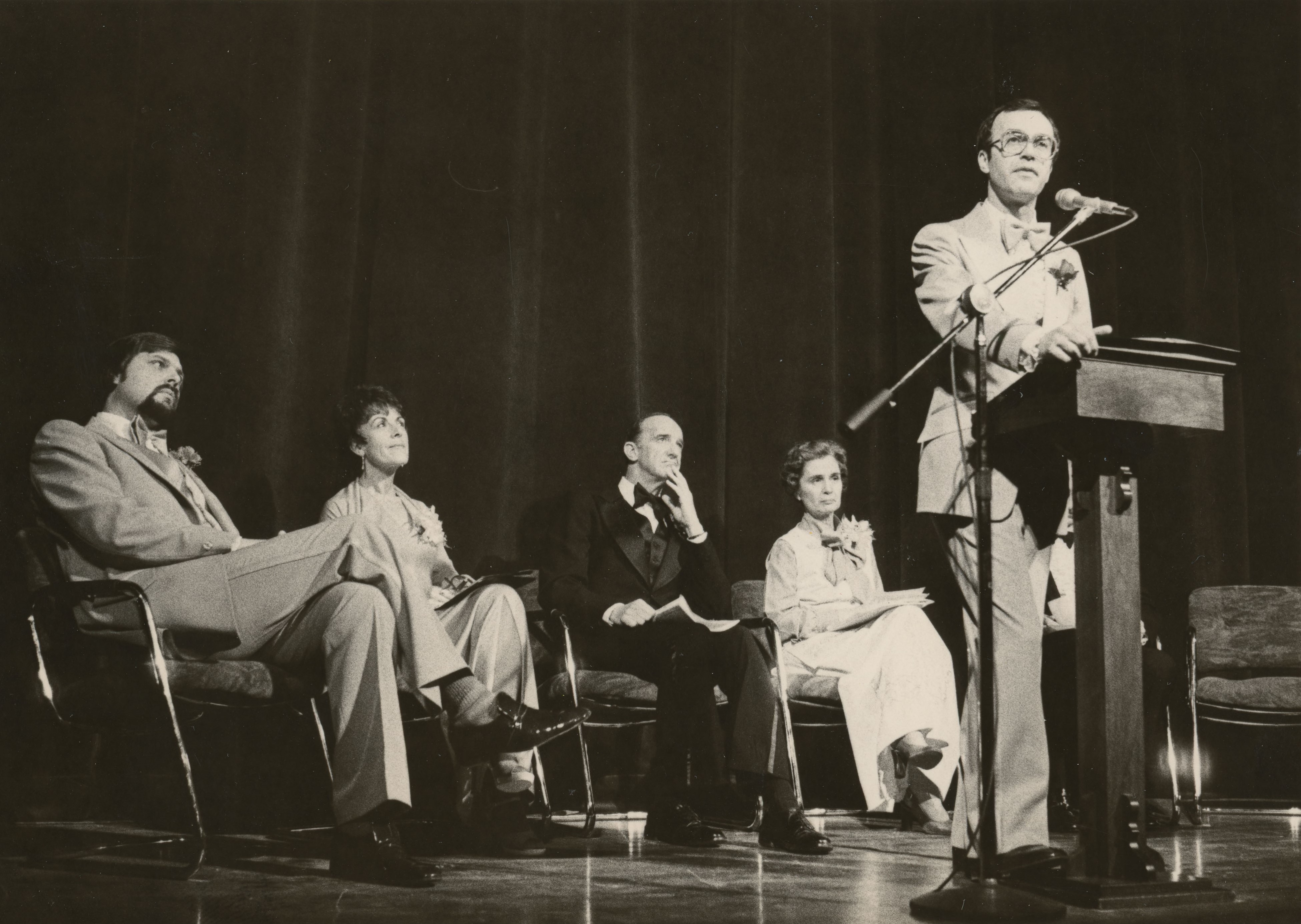 Man speaking on stage at a podium, five people seated in chairs behind him