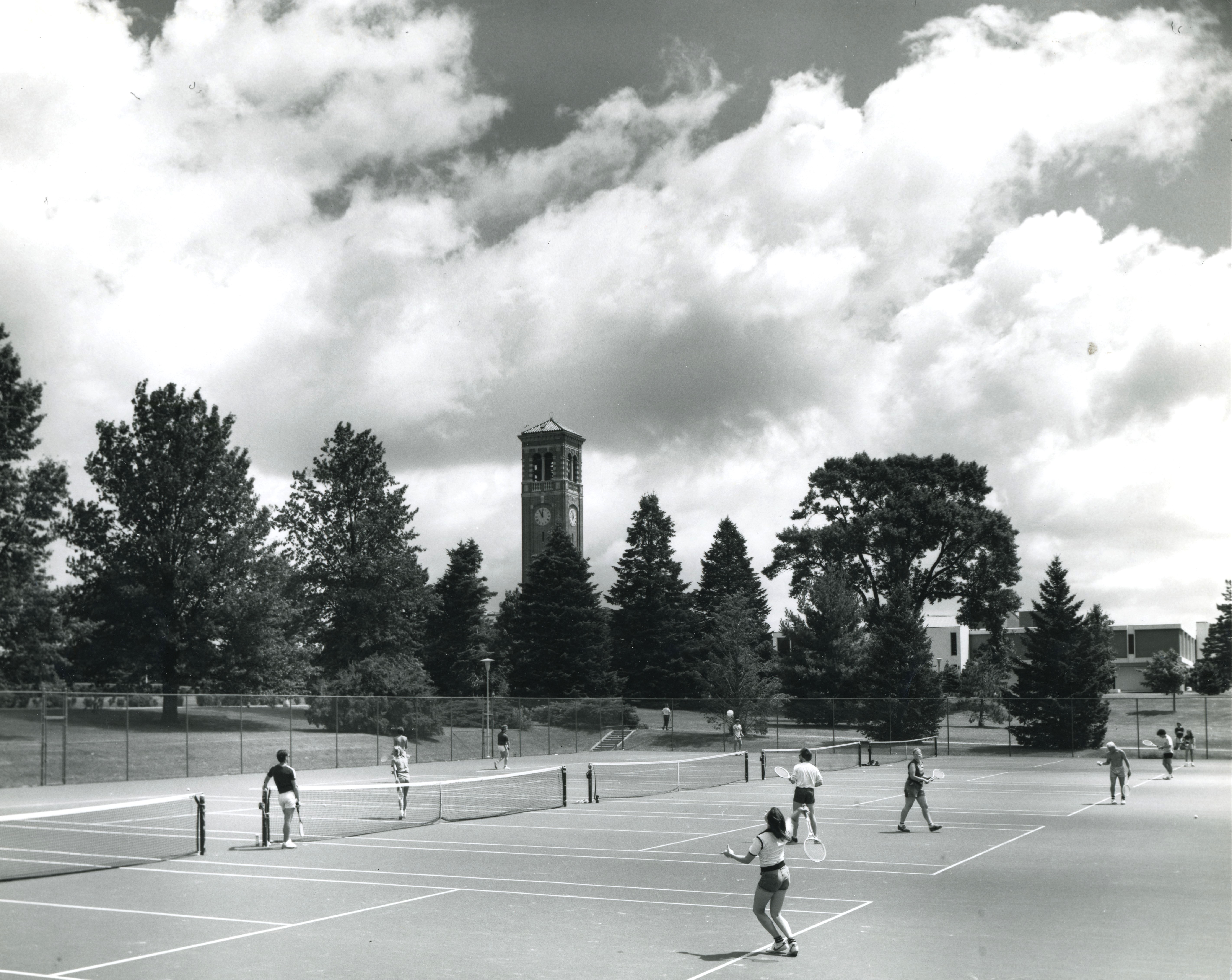 About 10 people playing tennis on four outdoor courts with the Campanile in the background