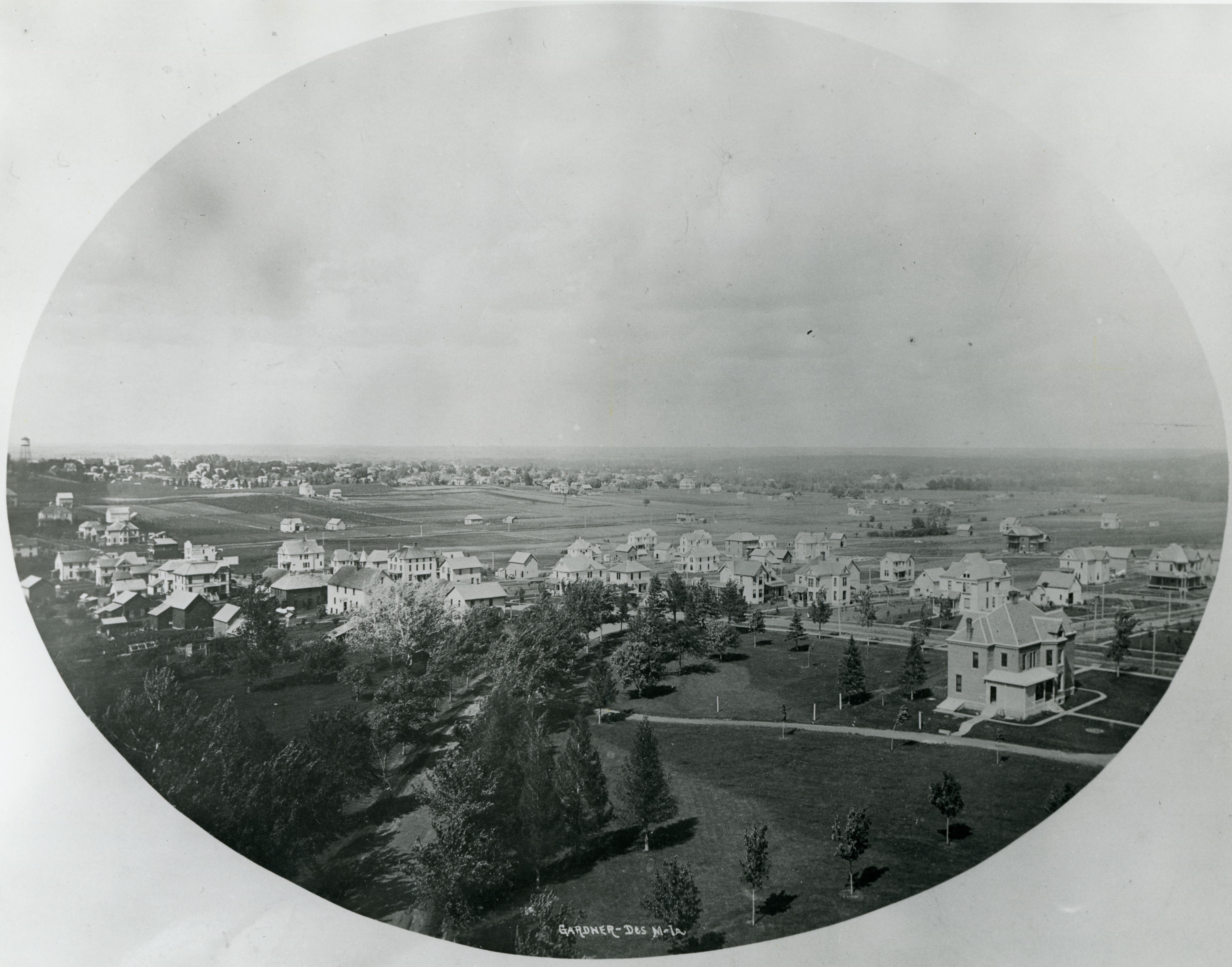 Aerial of a cluster of buildings along a street, scattered trees in the foreground, another larger cluster of buildings in the distance, caption reads Gardner- Des M - IA