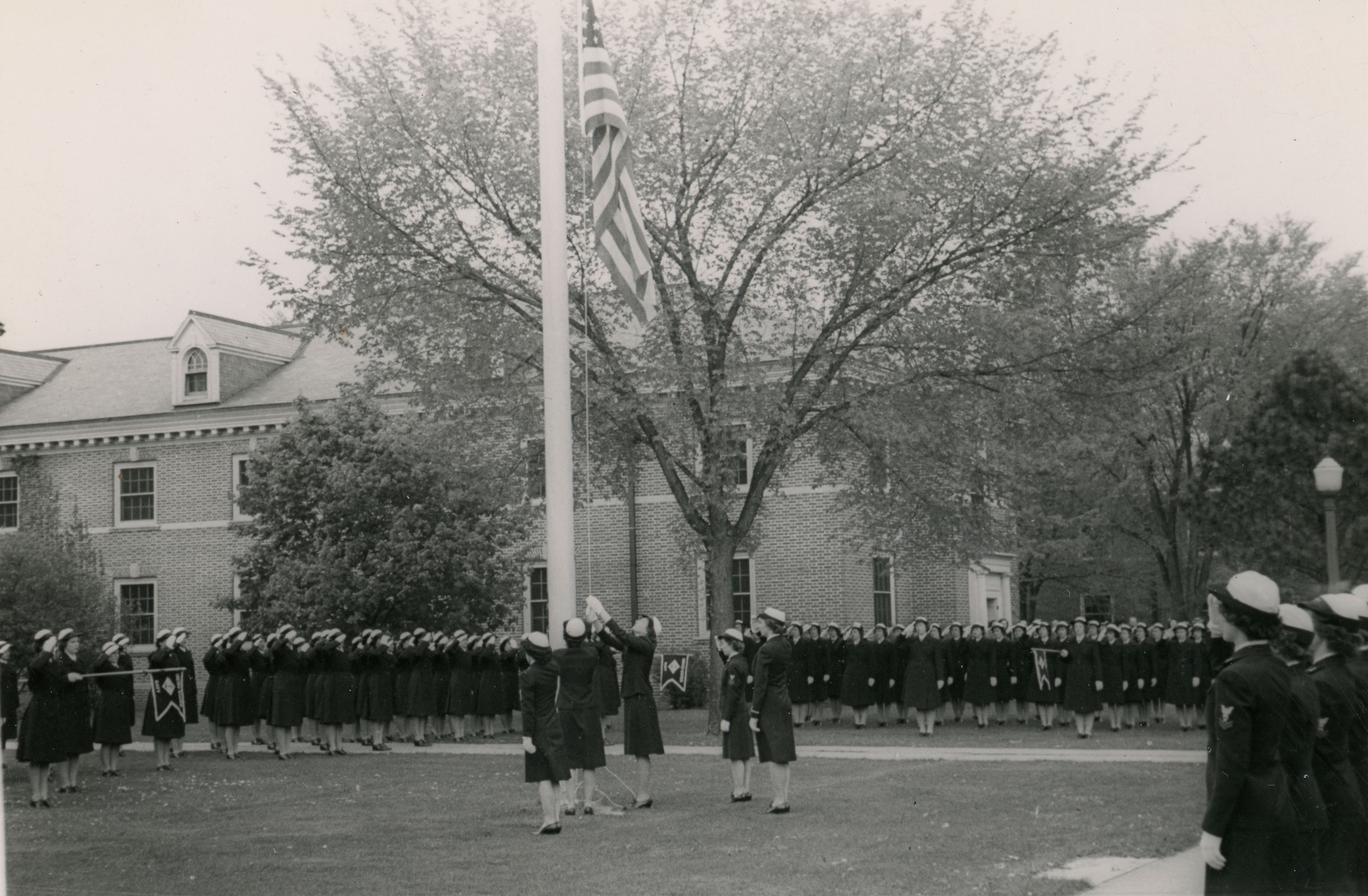 Five women raising a United States flag up a poll, others standing in rows, saluting and watching; all in military uniforms with dresses and caps