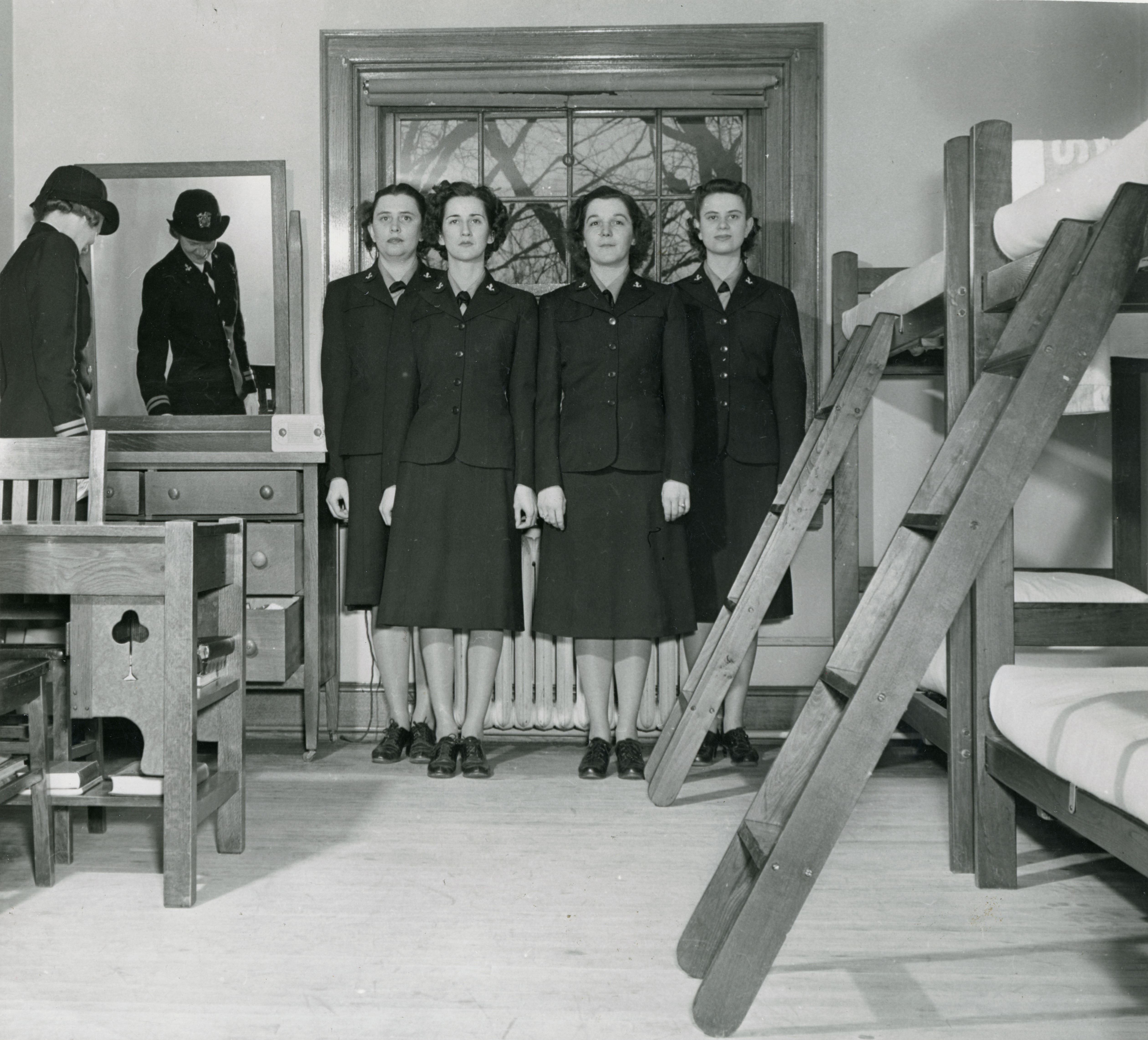 Four women in military dresses standing at attention against a window, one woman in a similar uniform with a cap inspecting the dresser to the left; bunk beds to right