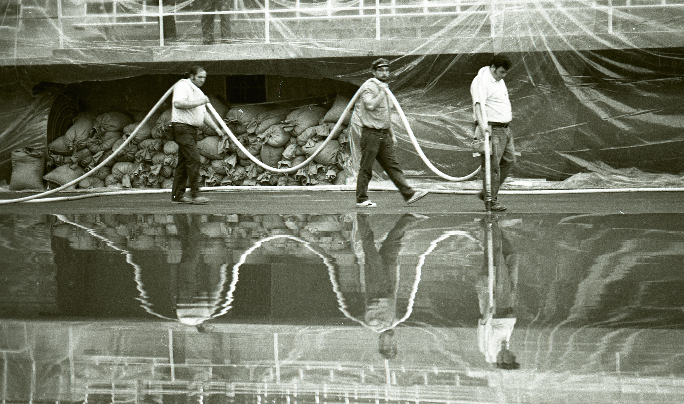 Three men carrying a hose; they are reflected in a pool of water on the floor, wall behind them is draped with plastic and piled with sandbags