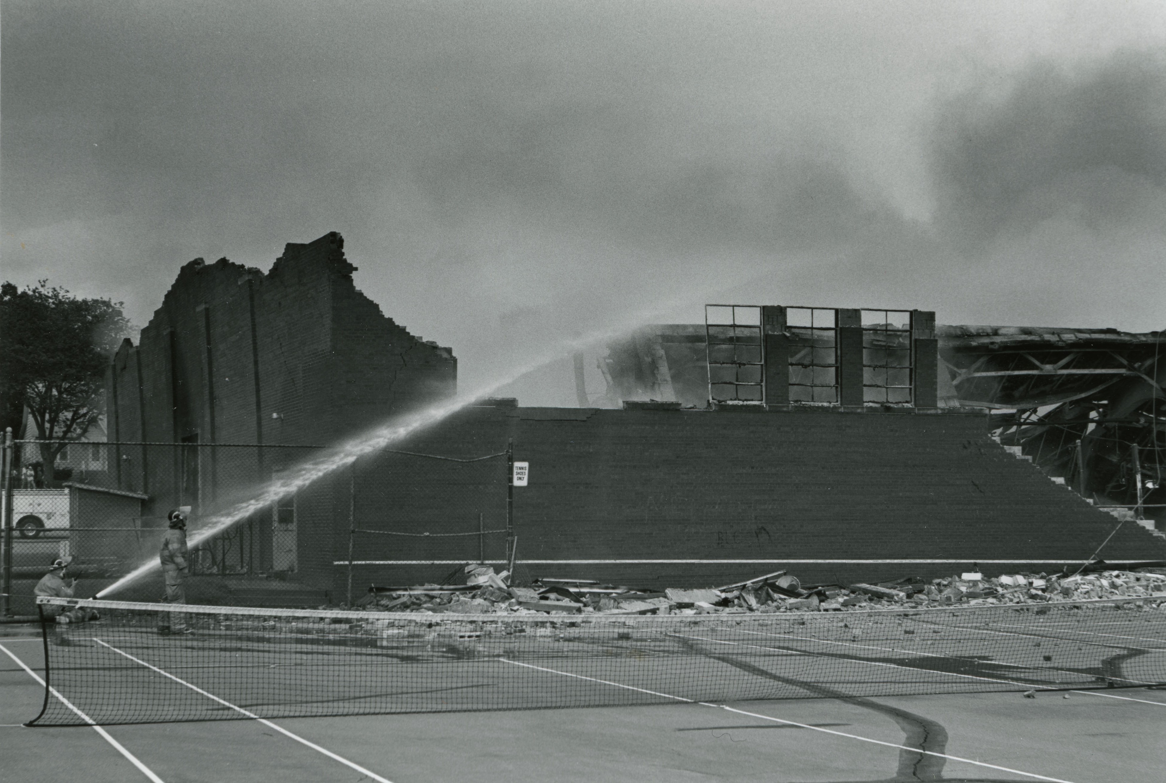 To left, two firefighters pointing a hose spraying water at a mostly-destroyed, still-smoking brick building; outer walls are somewhat standing, but roof and other walls are collapsed