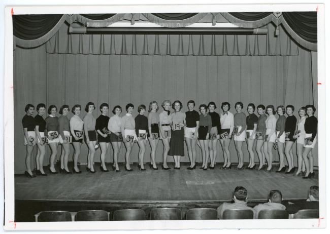 Several young women lined up on a stage for the Old Gold Beauties contest, each is holding a piece of paper with a number written on it 