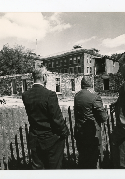 Five men behind fence observing remains of building after fire