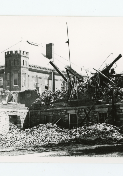 Fire-damaged building remains with collapsed bricks and timbers, intact building behind ruins