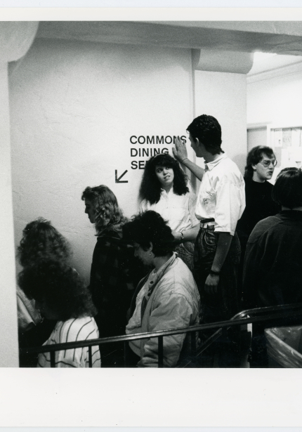Students conversing while waiting in a line to go downstairs, partially visible wall sign reads Commons Dining