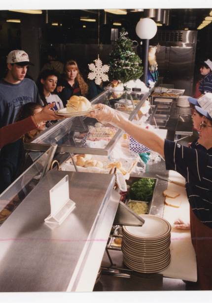 Employees on right side of cafeteria serving line fill plates and hand them to diners on other side