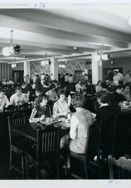 Groups of people seated at tables in dining area with food line along back wall