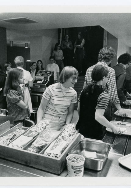 People grabbing silverware, bread, and trays in cafeteria after paying at register