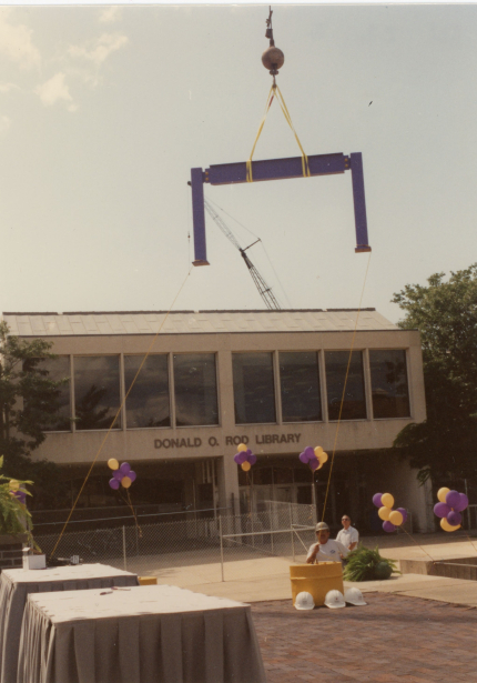 Crane lifting beam to roof of Donald O. Rod Library, platform and balloons in the foreground