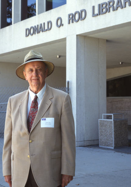Man in suit jacket and hat with nametag standing in front of building named Donald O. Rod Library