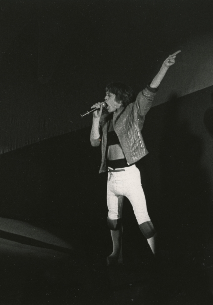 Man holding and singing into a microphone with one hand, other arm extended and pointing up and to the right