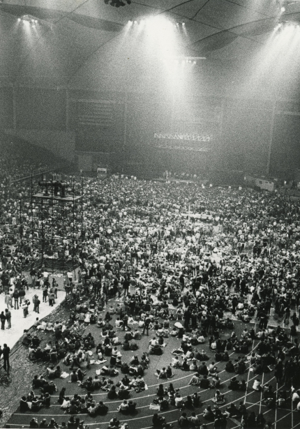 Indoor football field and stadium seats full of people, stage set up at one end