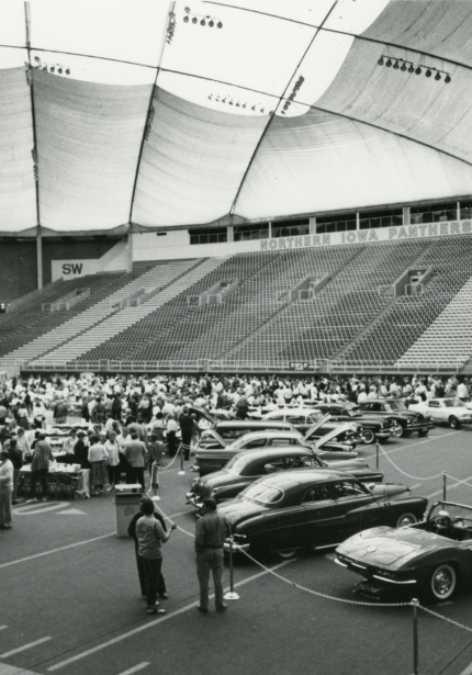 Car show on football field in UNI-Dome with crowd outside roped-off area where cars are parked