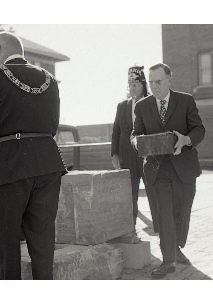 Man carrying a small box to a table, two men in Masonic regalia gathered around it and a reporter in the background