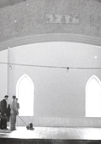 Four men working in room, three appear to be sanding a wooden stage at the front, upright piano to right and grand piano to left