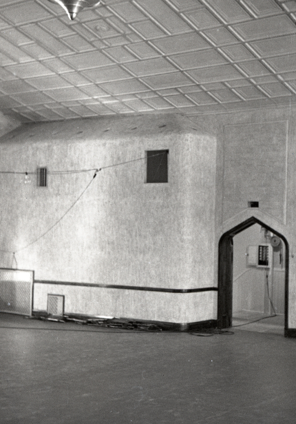 Room with bare floor and supplies piled against the wall, makeshift lights strung across the ceiling and an open arched doorway to the right