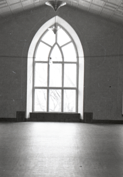 Open room with larged pointed arched window flanked by two smaller windows to left and right, bare floor, and lights strung on a wire across the room