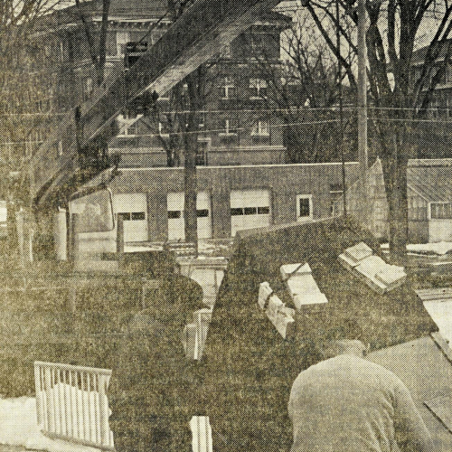 Printed in newspaper, two men steadying a piece of equipment cabled to a crane
