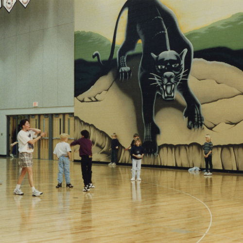 Teacher and about twelve students playing in a gymnasium with a mural of a panther on one wall