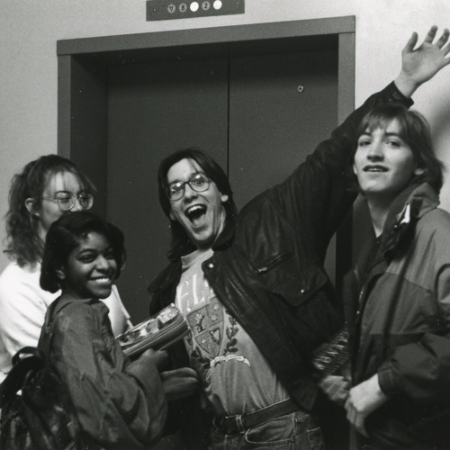 Four smiling high school students standing in front of an elevator, one posing with arms extended