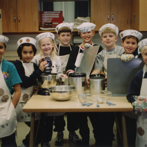 Eight third graders in aprons, hats, and oven mitts gathered around a kitchen table with baking utensils, smiling at the camera